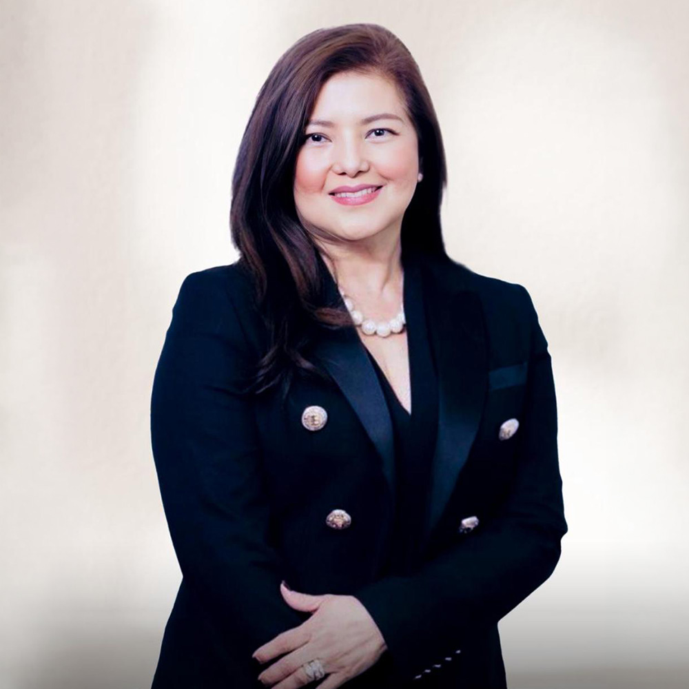 Smiling woman with long hair in a black blazer and pearl necklace on a light background.
