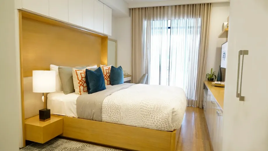 Bright master bedroom with wooden bed frame, patterned pillows, lamp, and sheer curtains on window.