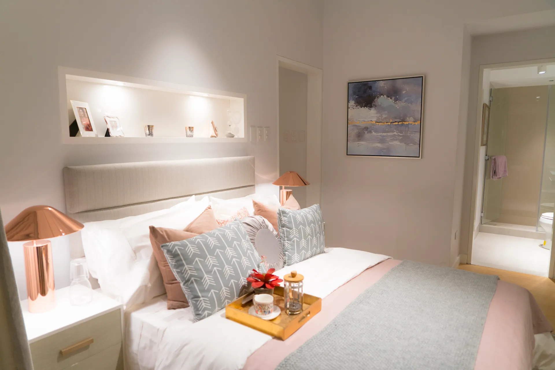Cozy bedroom with white and pastel bedding, patterned pillows, rose gold lamps, and a tray with cup and flower.