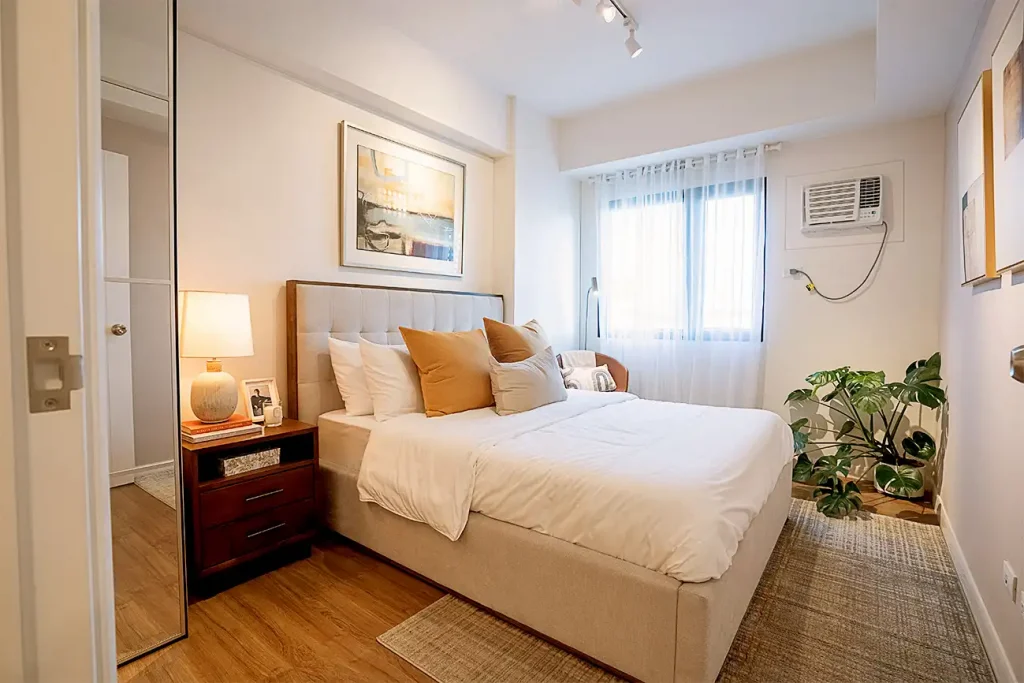 Cozy bedroom with beige bed, white bedding, brown pillows, wooden nightstand, and plant near window.