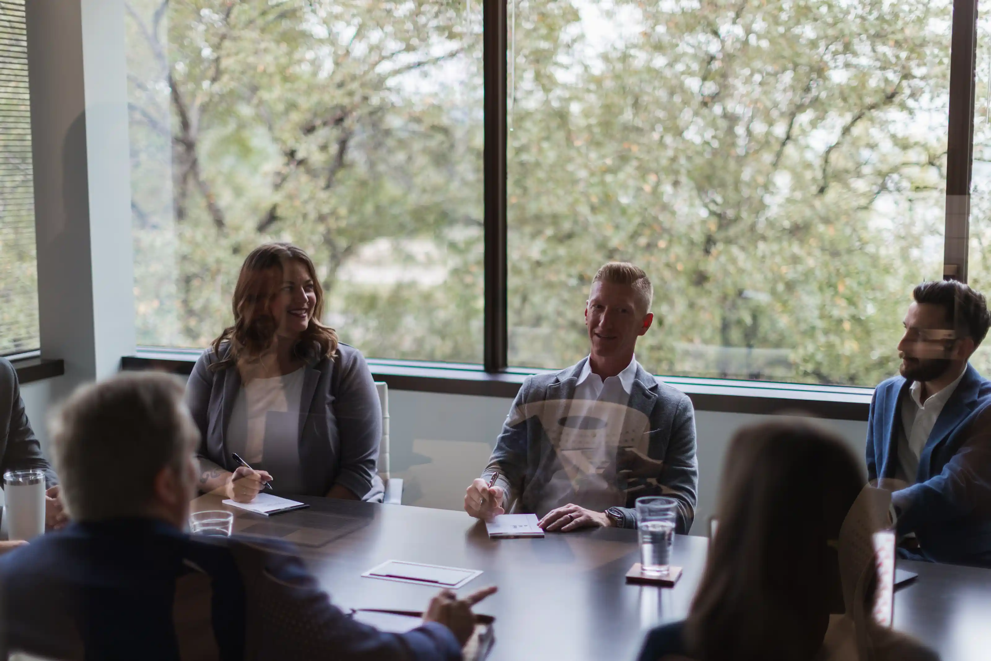 Group of five people sitting around a conference table in a modern office with large windows and trees outside, engaged in a meeting, with two people taking notes and others listening attentively.