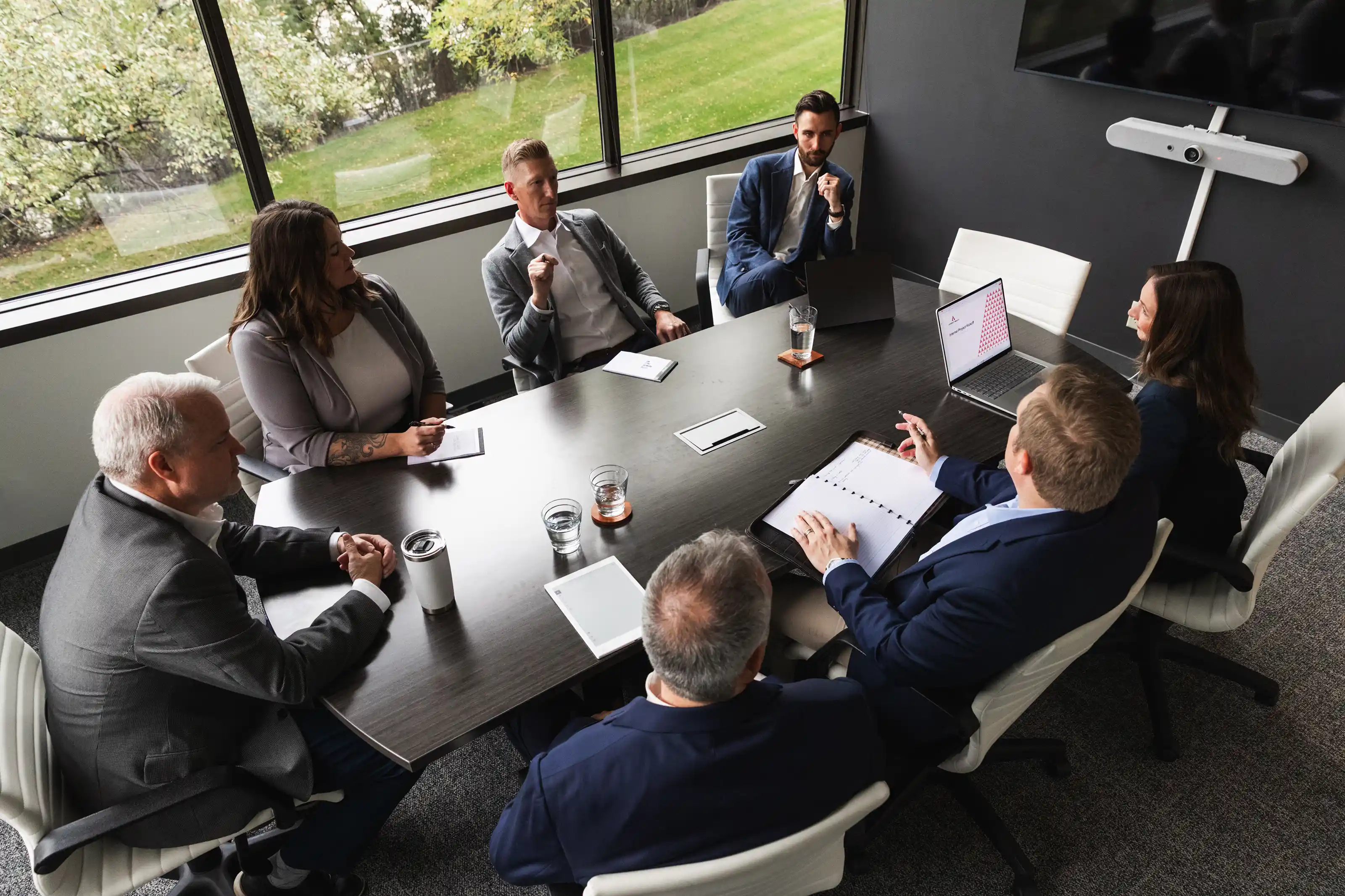 Seven business professionals seated around a dark wooden conference table in a modern office with large windows, engaged in a meeting; some have laptops, notepads, and documents, while others listen attentively.