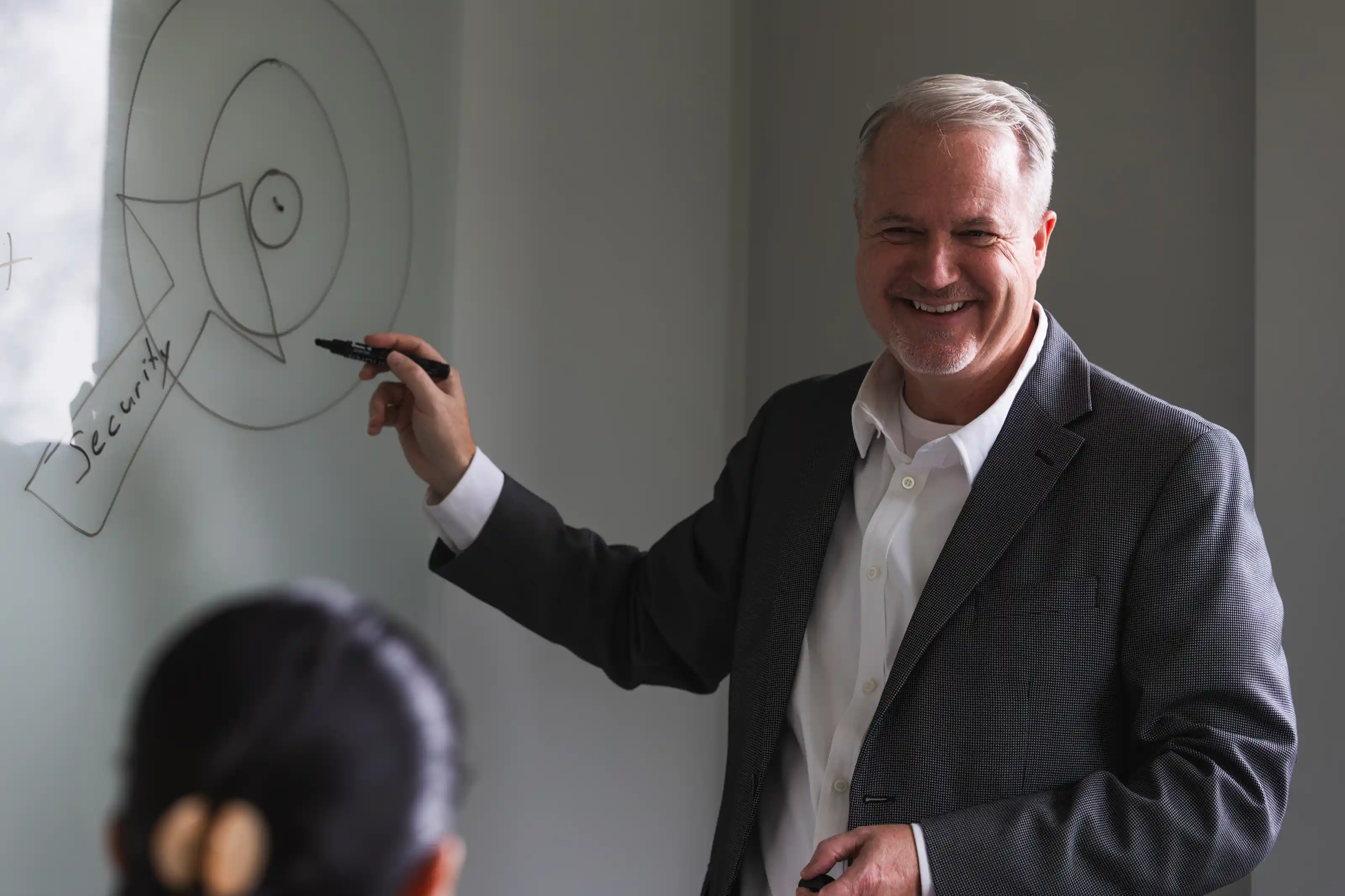 Smiling mature man in a suit holding a marker and pointing to a diagram with concentric circles and an arrow labeled 'Security' on a whiteboard, with a blurred person facing him in the foreground.