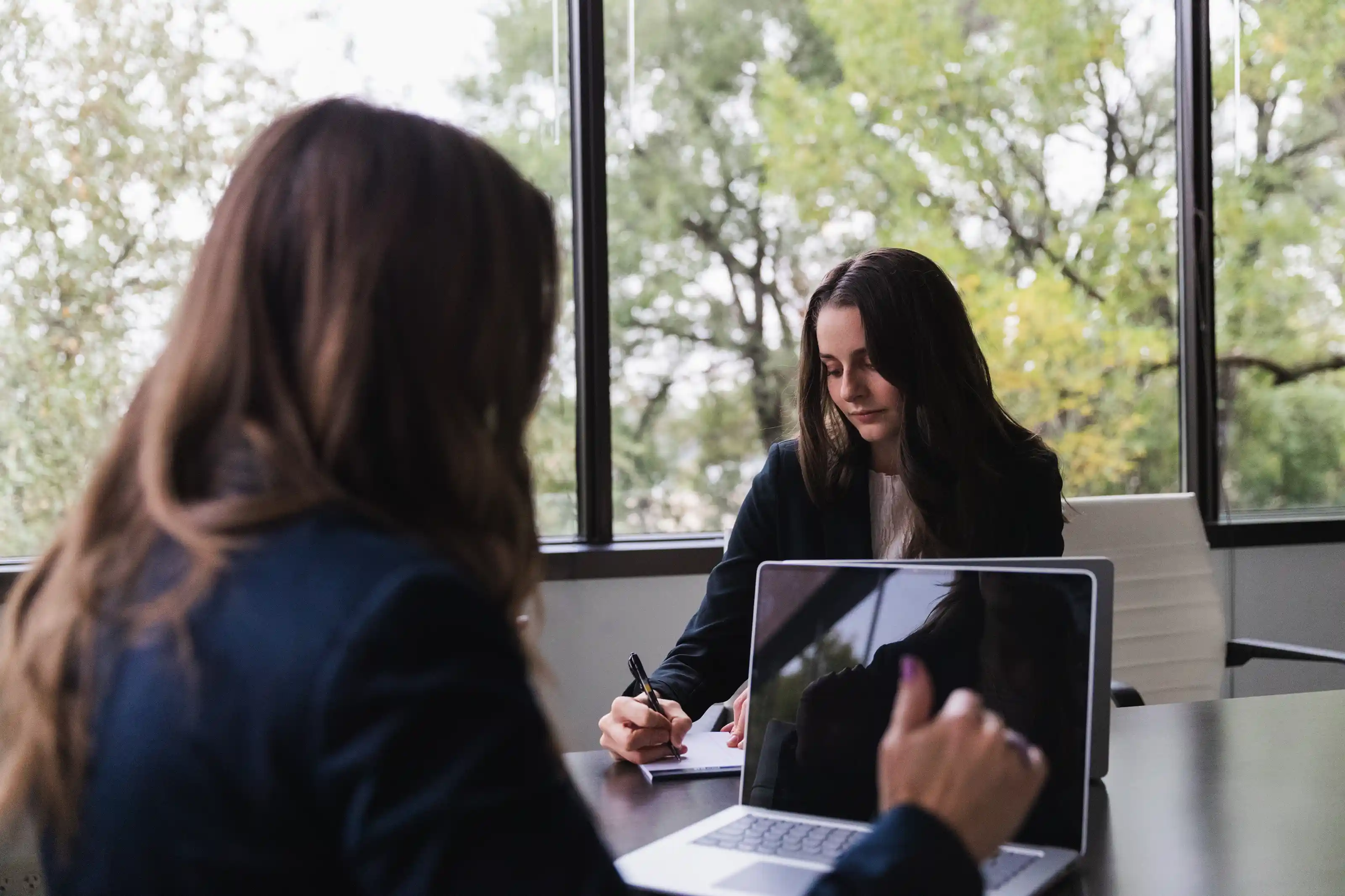 Two women in a modern office, one writing on paper while the other uses a laptop with a reflective screen, large windows showing green trees outside.