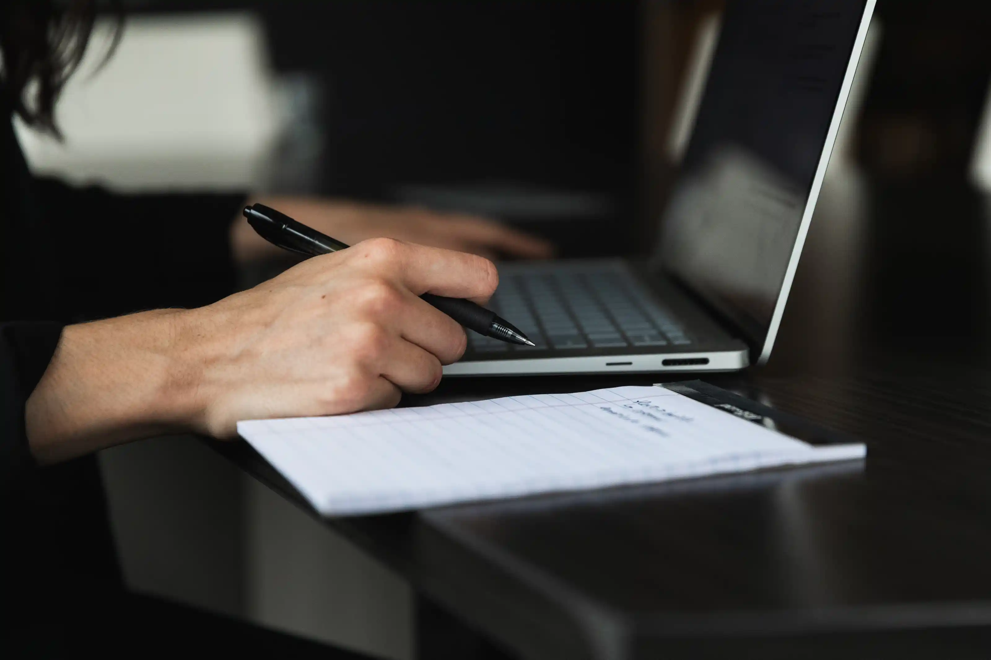 Close-up of a person's hand holding a black pen over a lined notepad on a dark desk, with a laptop keyboard visible in the background.