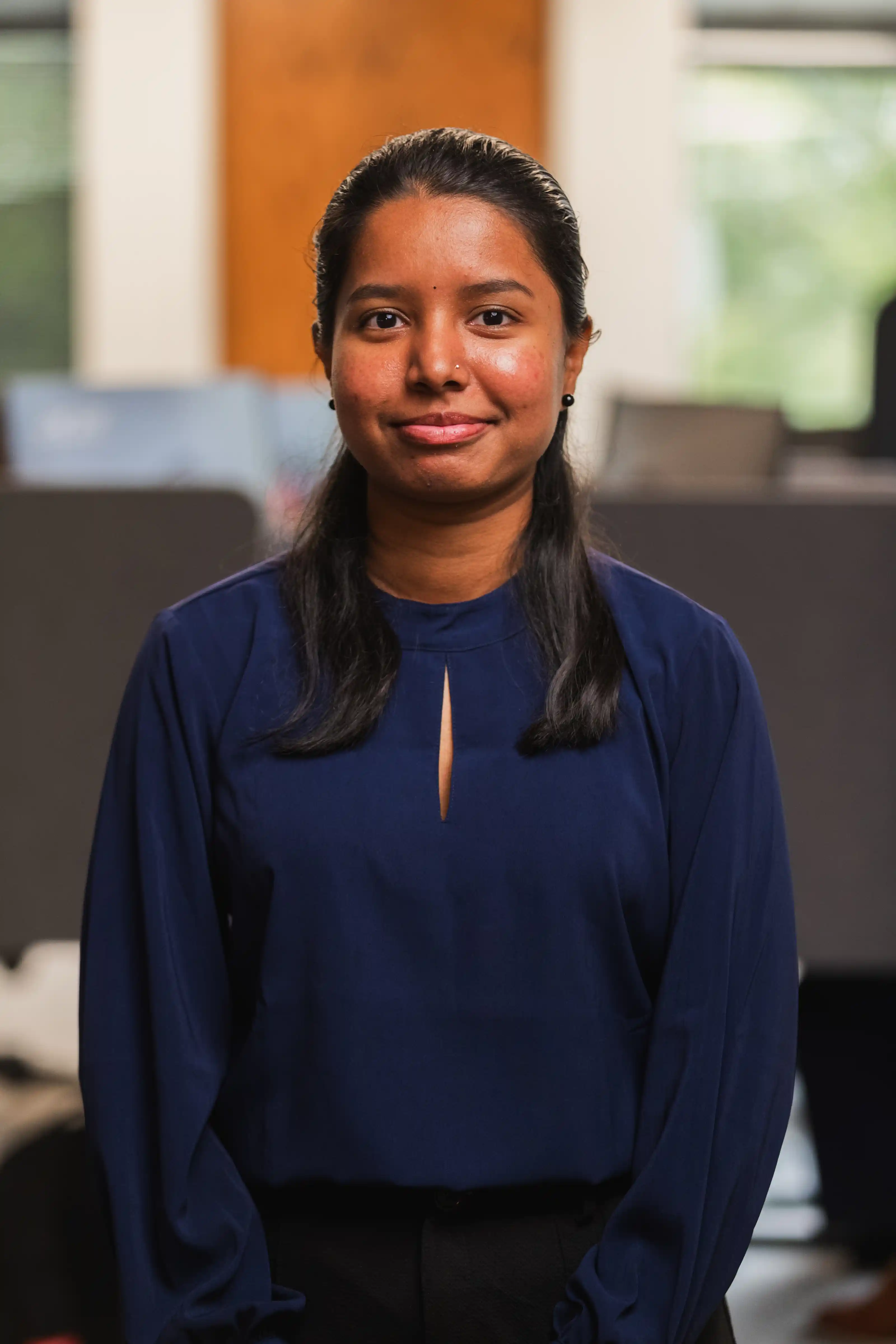 A woman with long black hair, wearing a blue blouse with a small neckline slit, stands indoors with a neutral expression and a blurred office background.