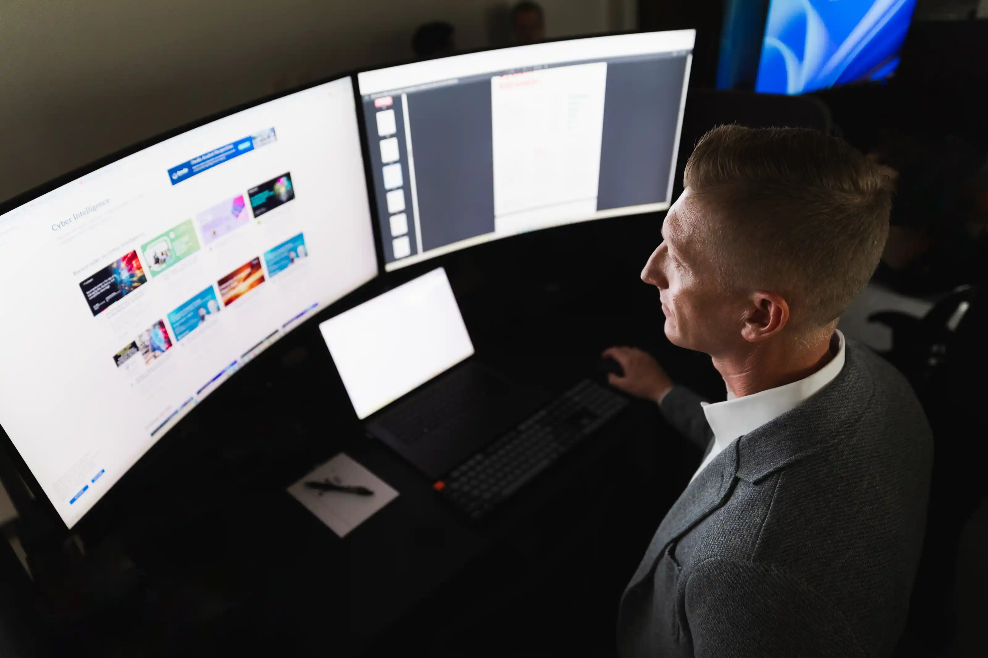 Man in a gray blazer working at a dark desk with a keyboard, laptop, and two large curved monitors displaying colorful digital content and documents.