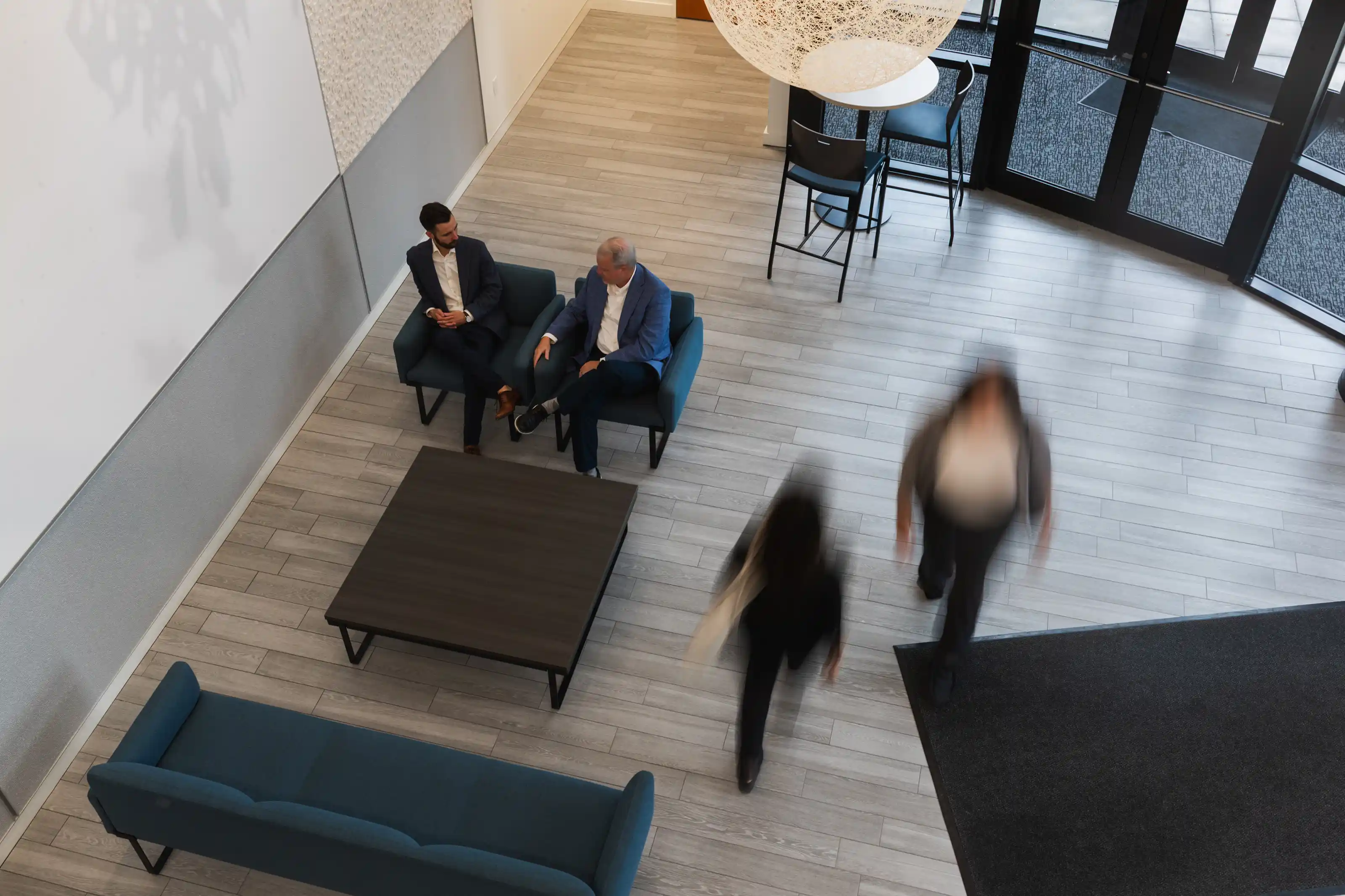 Overhead view of an office lobby with two men sitting and talking on blue armchairs around a dark coffee table, while two blurred women walk past near glass entrance doors.