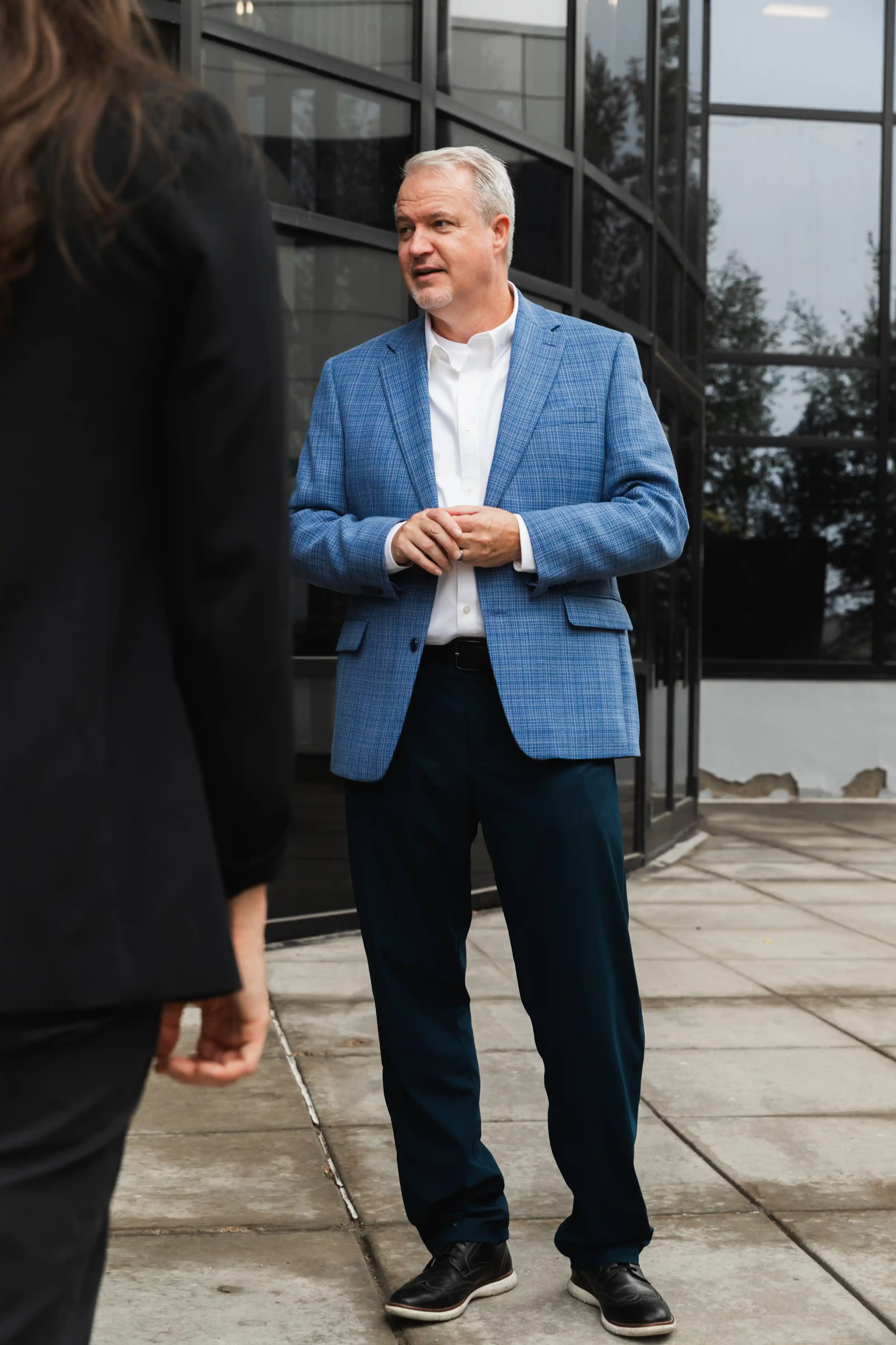 An older man with gray hair wearing a blue checked blazer, white shirt, dark pants, and black shoes stands on a sidewalk outside a modern glass building, looking slightly to his left and clasping his hands.
