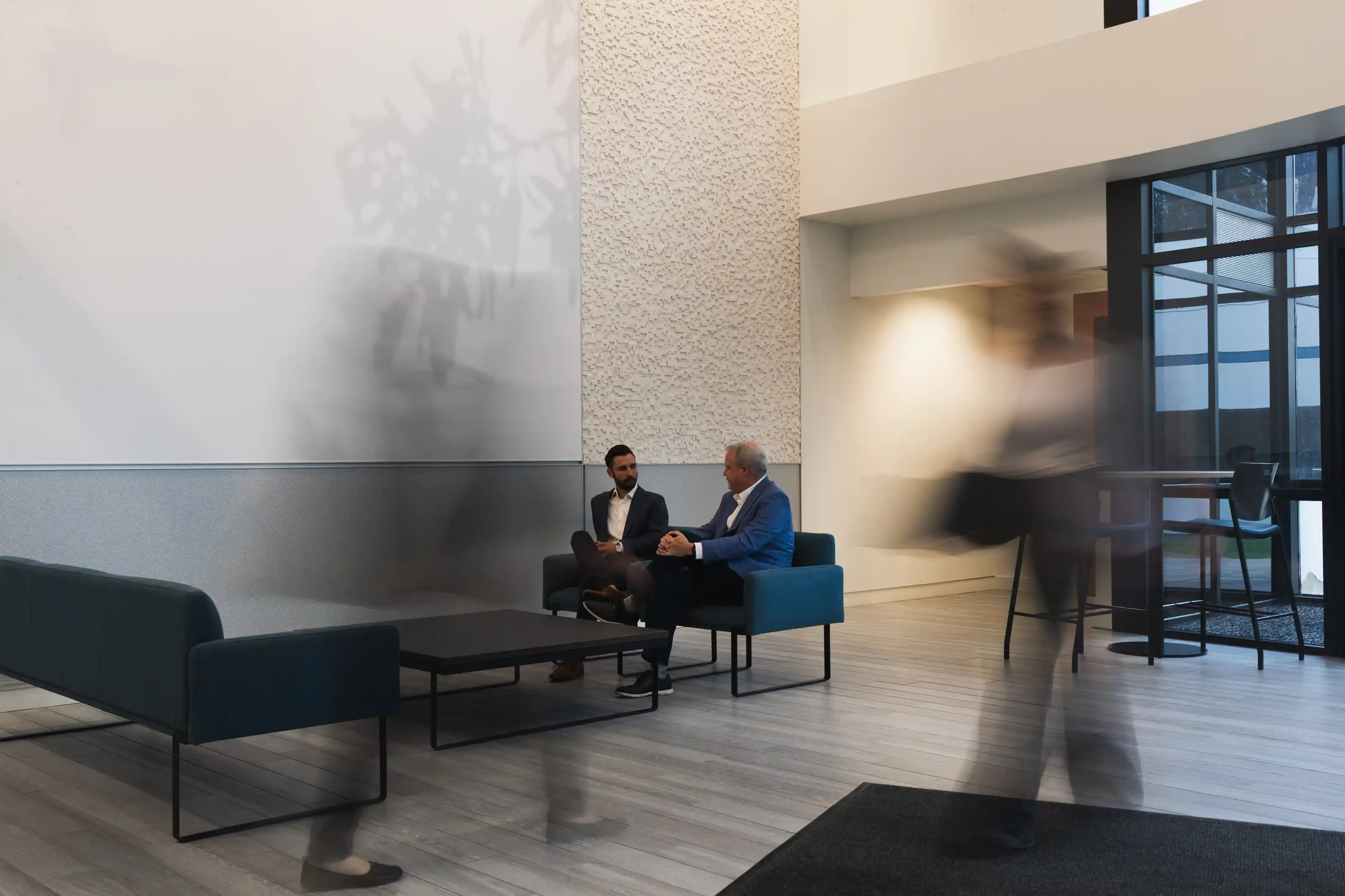 Two men sitting and talking in a modern office lobby with minimalist furniture, while blurred figures move around them.