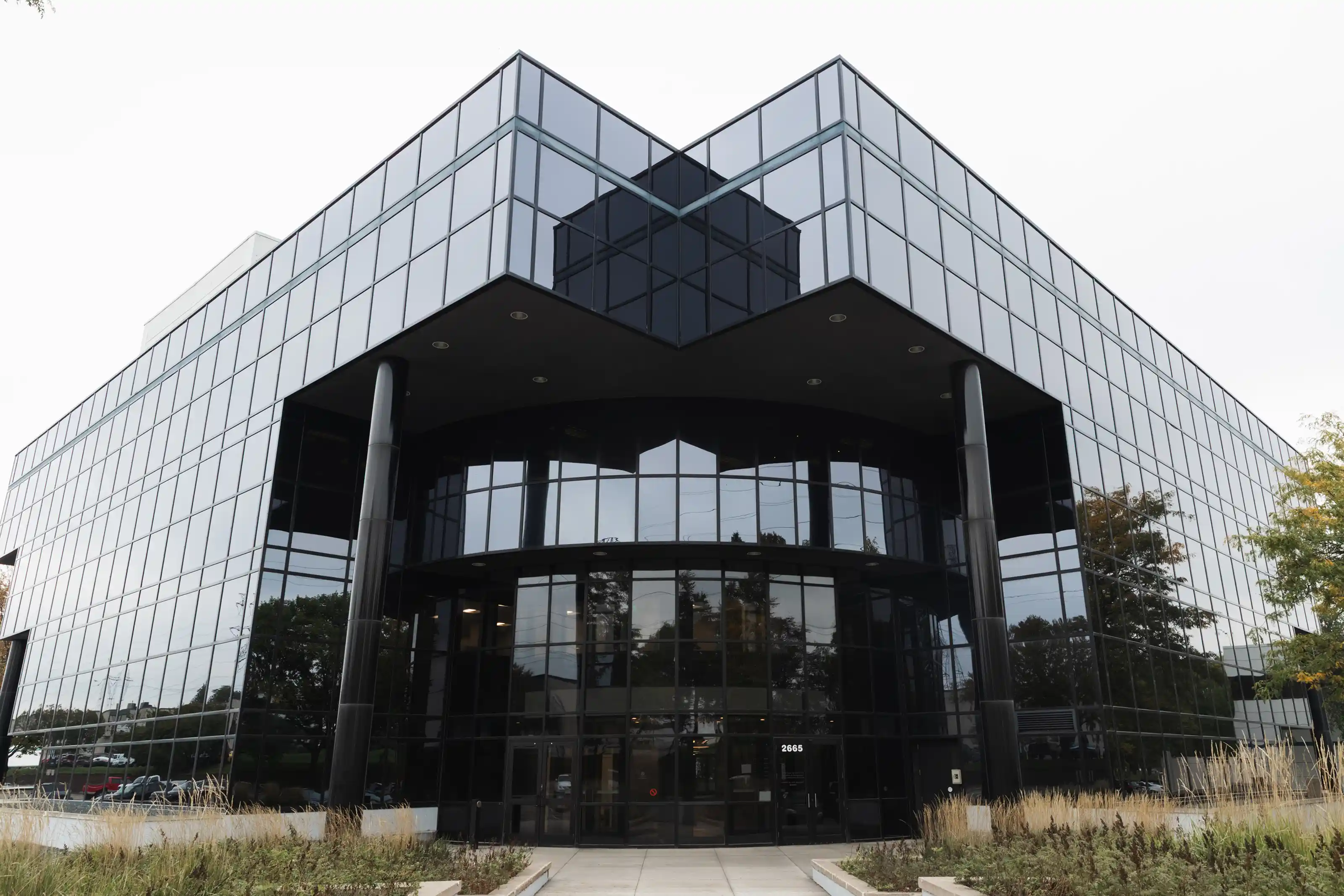 Modern office building with reflective glass windows and two large black columns flanking the main entrance at 2665, surrounded by plants and trees.