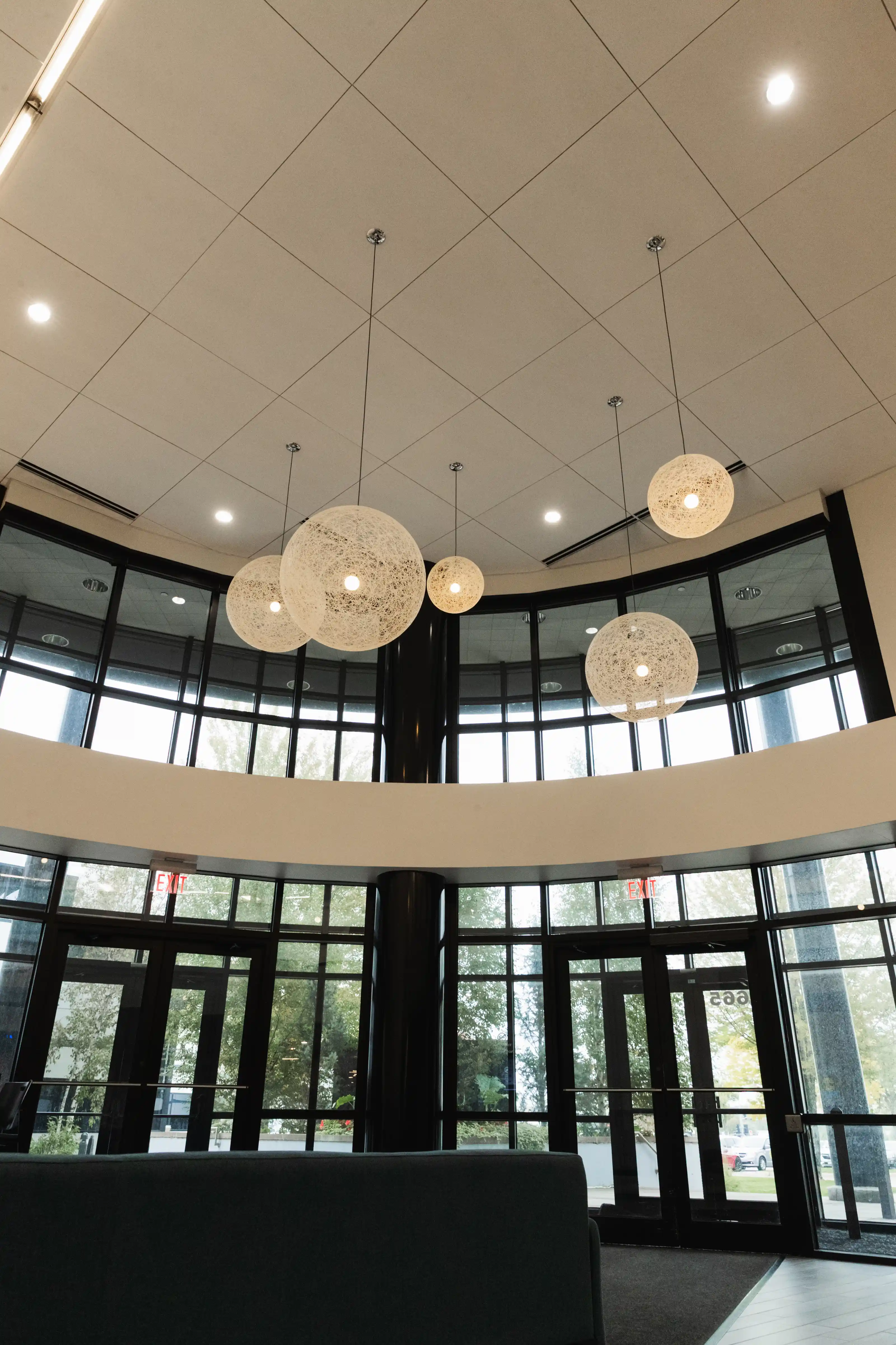 Interior view of a modern lobby with large floor-to-ceiling windows and black-framed glass doors, showing trees outside; six spherical pendant lights with white woven designs hang from the ceiling above a dark seating area.