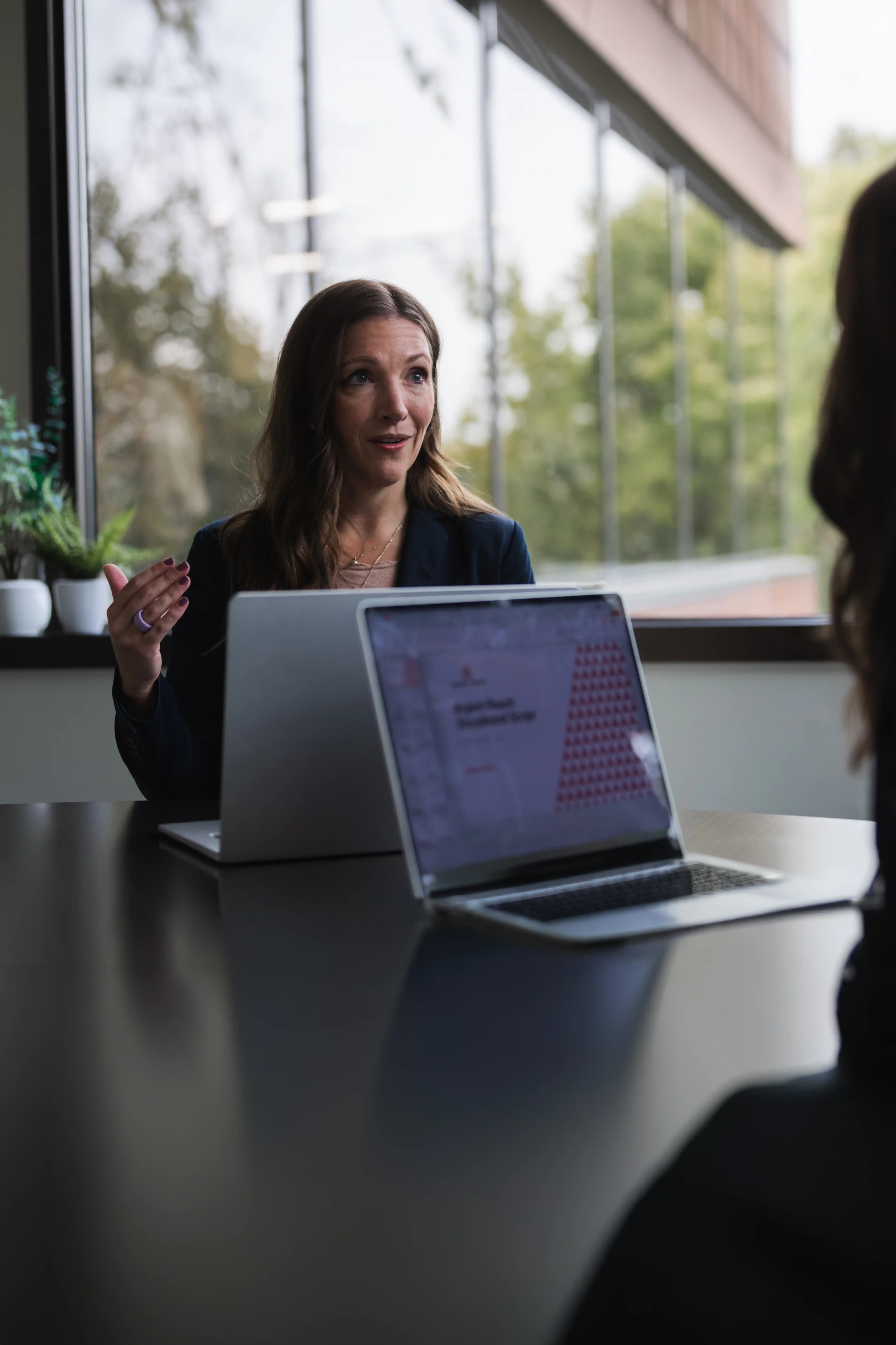 Two women in a meeting room with large windows; one woman is speaking and gesturing with her hand, seated behind a laptop, while the other is partially visible, also with a laptop open on the table.