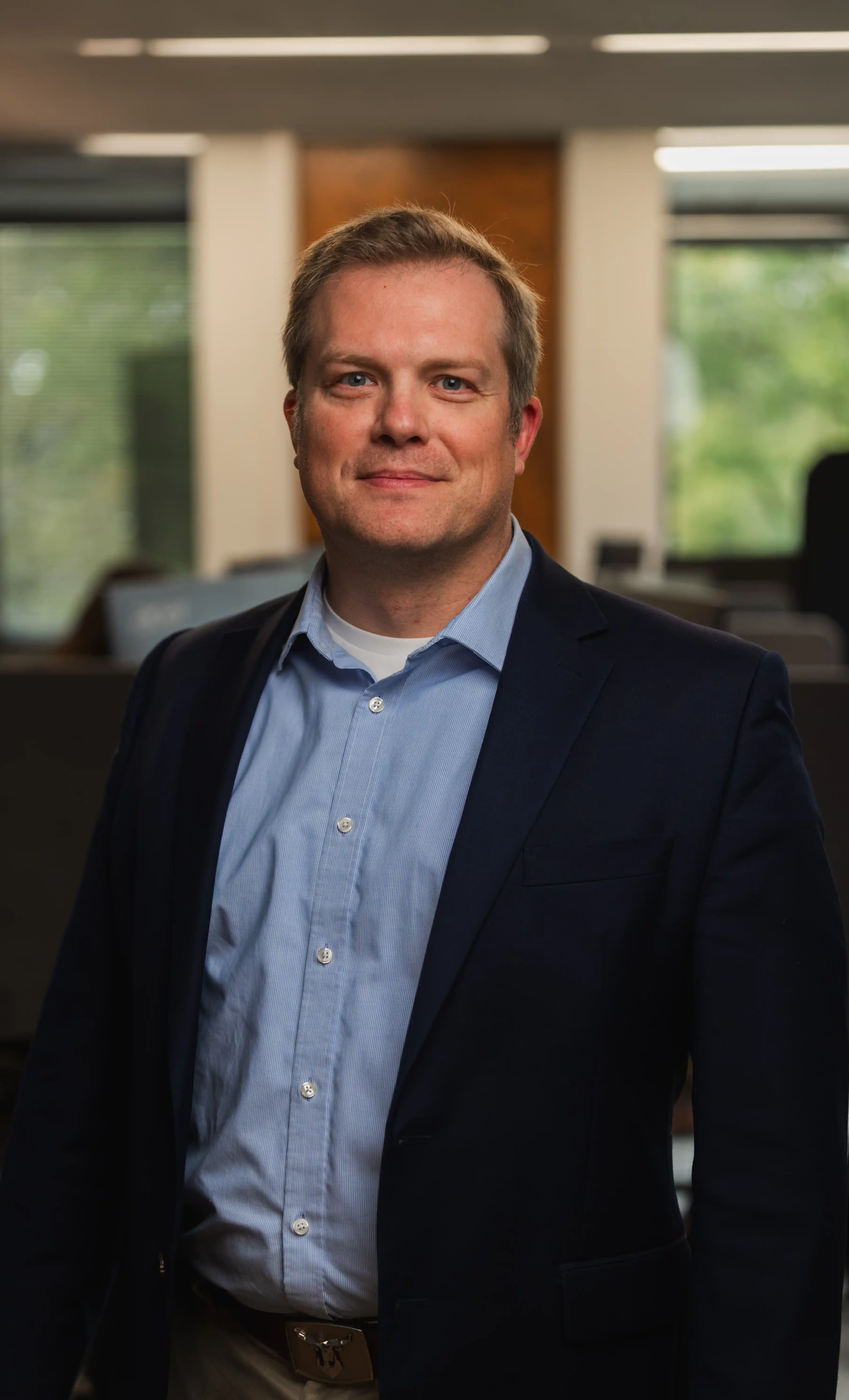 Portrait of a smiling middle-aged man wearing a light blue button-up shirt, a dark blazer, and beige pants standing in an office setting with blurred windows and desks in the background.