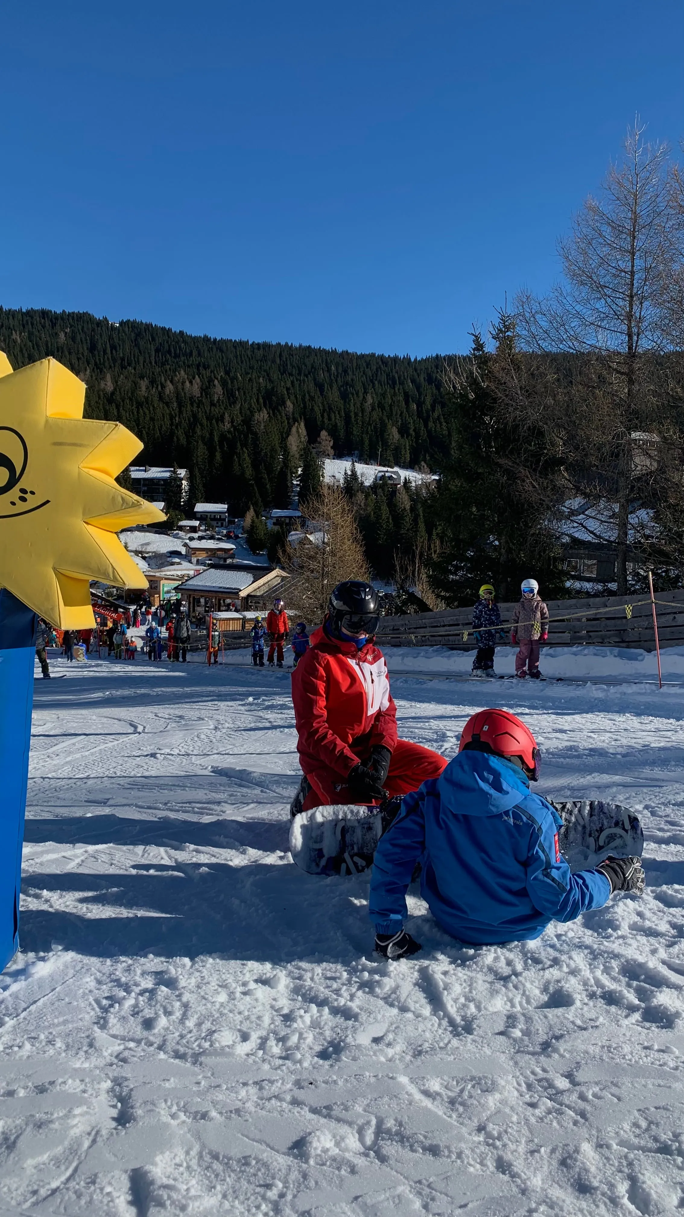 Instructor in red ski suit guides seated snowboarder in blue jacket on snowy slope with forest behind.