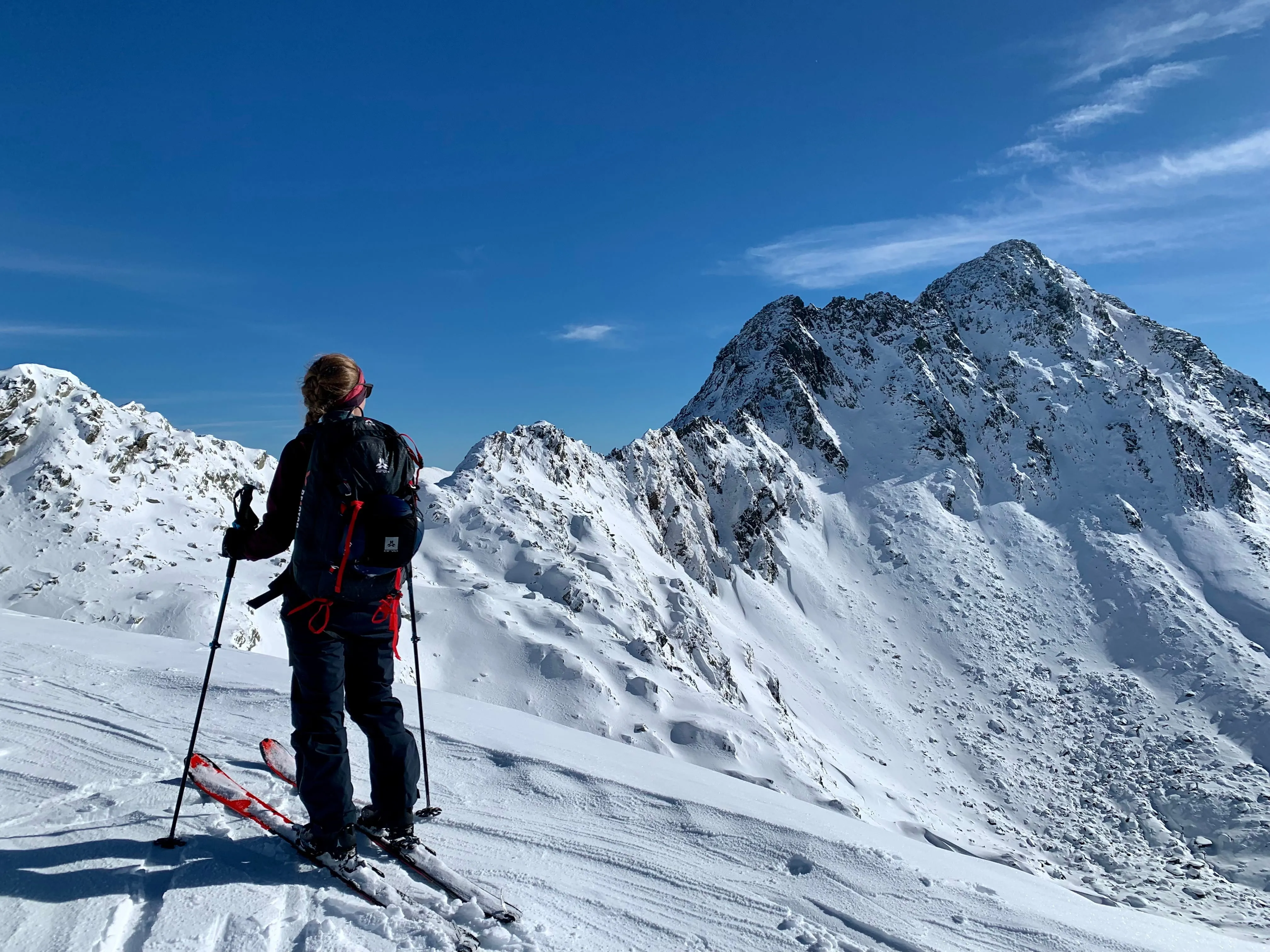 Skier with backpack stands on snowy slope looking at sunlit rugged mountain peak under a blue sky.