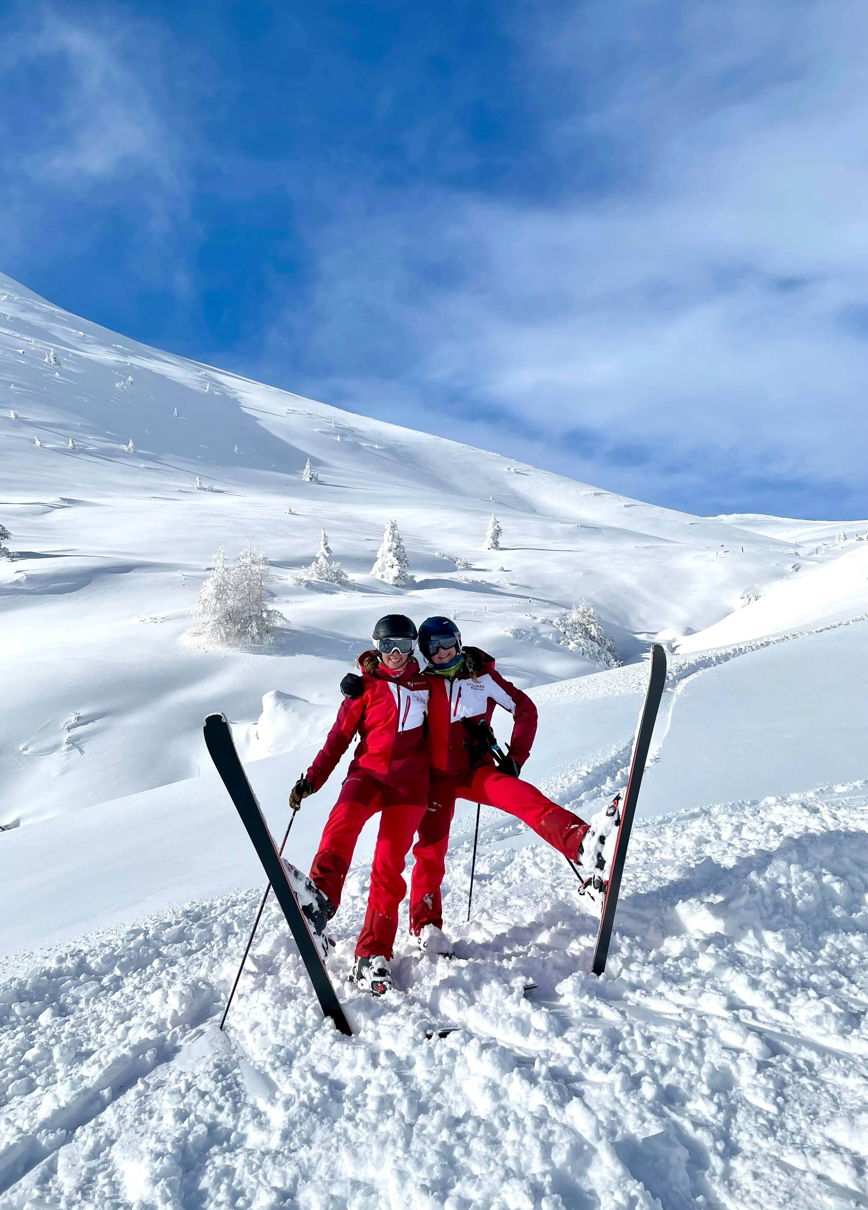 Two skiers in red jackets and helmets posing with skis raised on snowy mountain slope under blue sky.