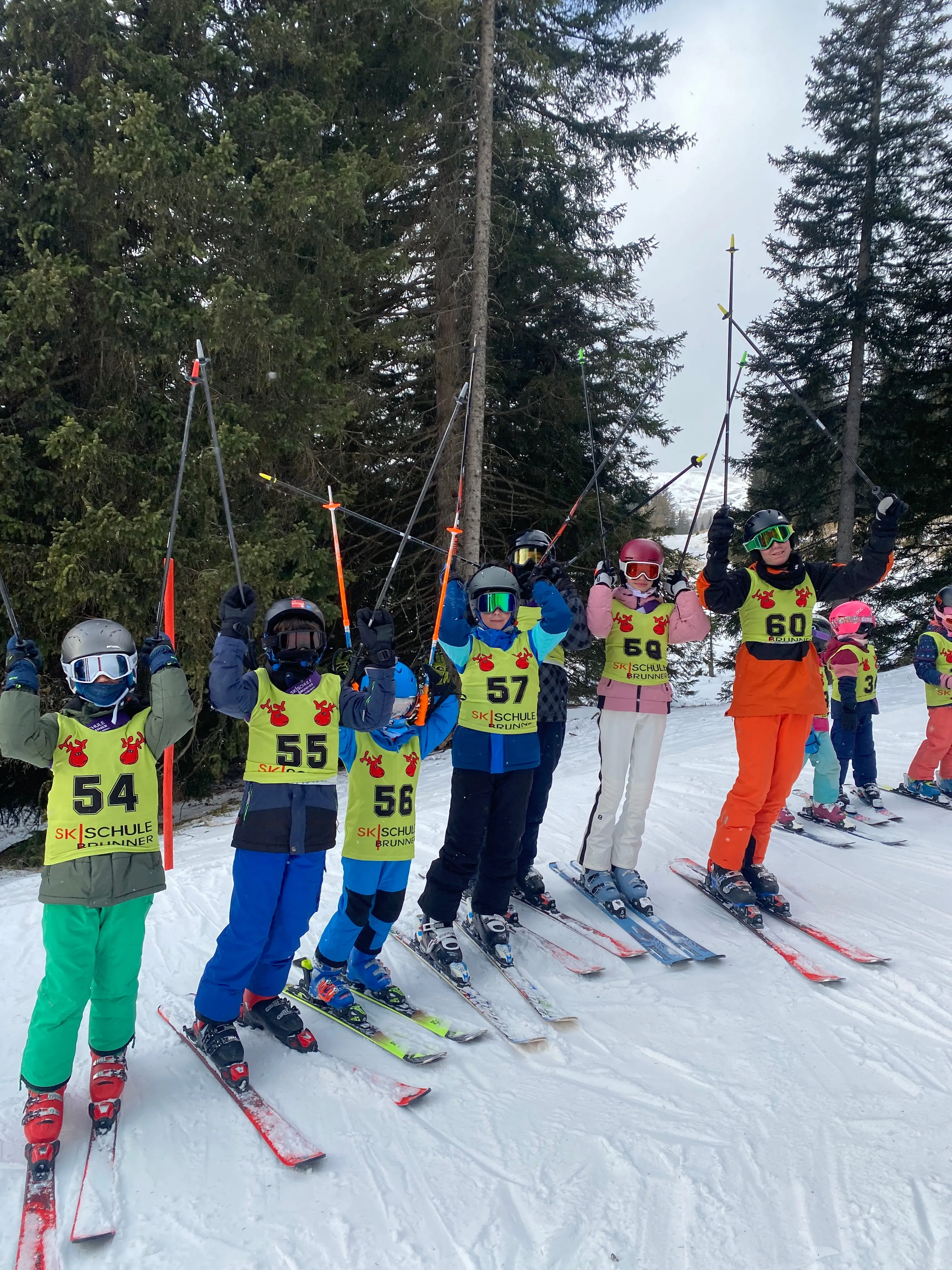 Children in numbered vests raising ski poles while standing on skis in snowy forest ski school.