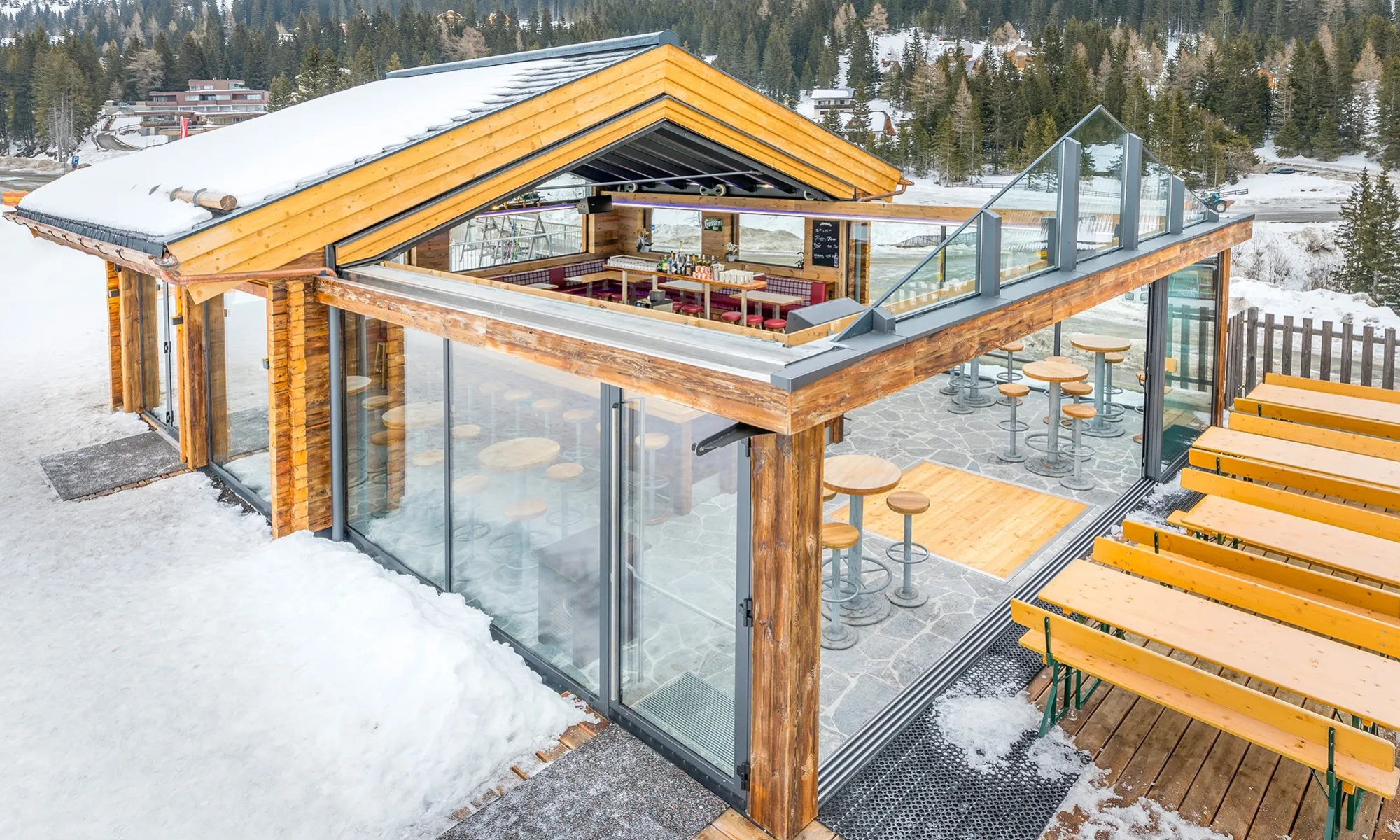 Wood and glass ski lodge bar with stools and outdoor seating surrounded by snow and pine trees.