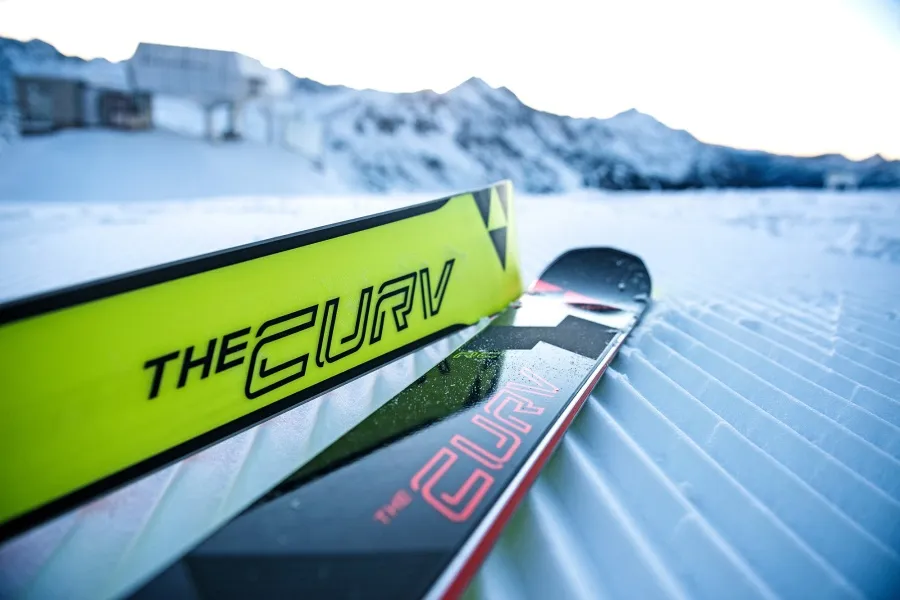 Zwei Skier mit der Aufschrift "THE CURV" liegen auf präpariertem Schnee vor Bergen.