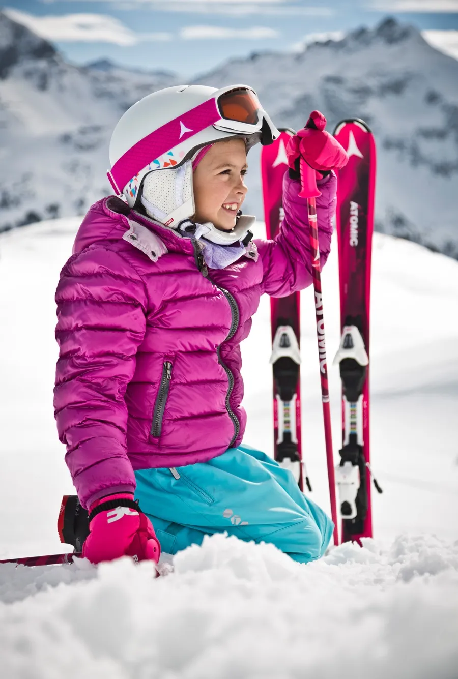 Lächeldes Kind in rosa Jacke und Helm kniet im Schnee mit Skiern und Stöcken im Hintergrund.
