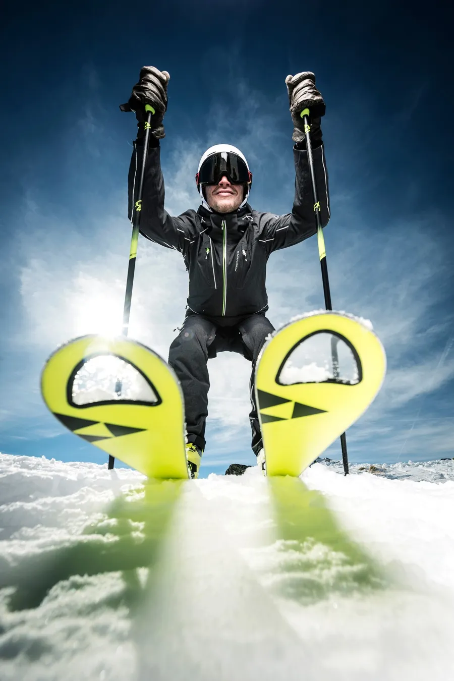 Skifahrer in schwarzer Kleidung und weißem Helm posiert auf leuchtend gelben Skiern im Schnee.