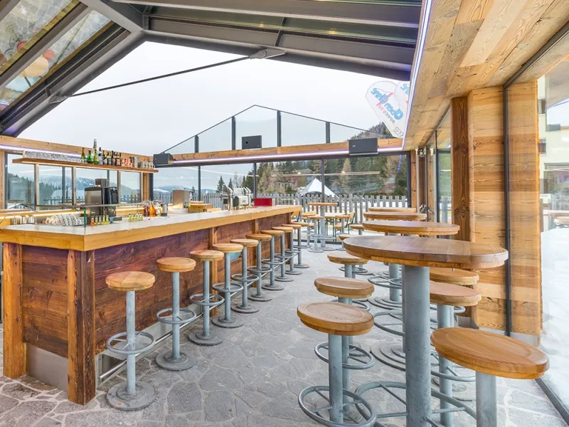 Helle Skibar mit Holztheke, hohen Barhockern und Tischen, Glaswänden zeigen verschneite Landschaft.