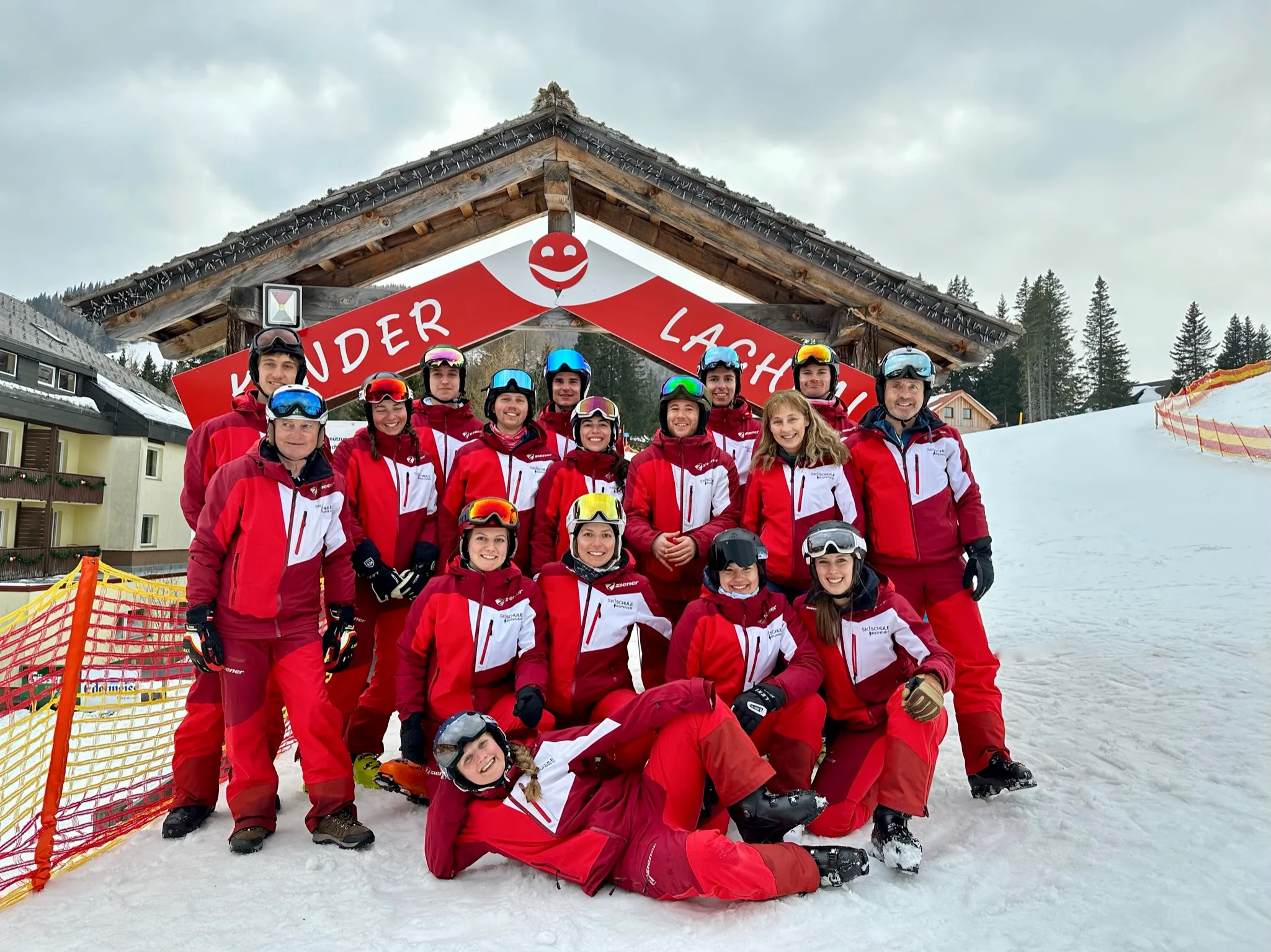 Gruppe von 16 Skilehrern in roten Skianzügen posiert vor einem Holzrahmen mit der Aufschrift Kinder Lachen.