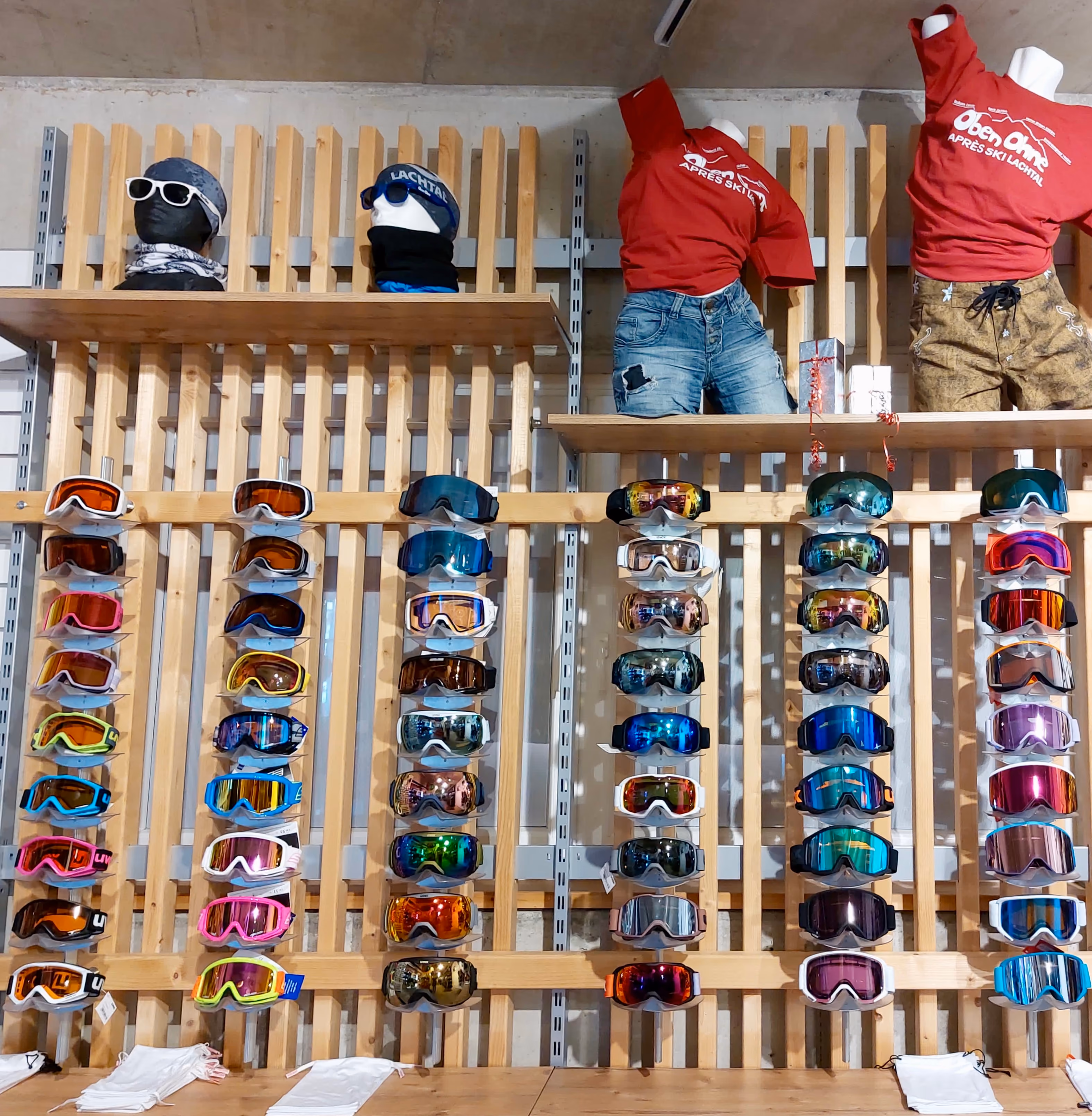 Verschiedene Ski- und Snowboardbrillen in bunten Farben an einer Holzlattenwand hängend, darüber zwei Schaumstoffköpfe mit Mützen und Brillen sowie zwei Schaufensterpuppen mit roten Après-Ski-Pullovern und Jeans oder braunen Hosen.