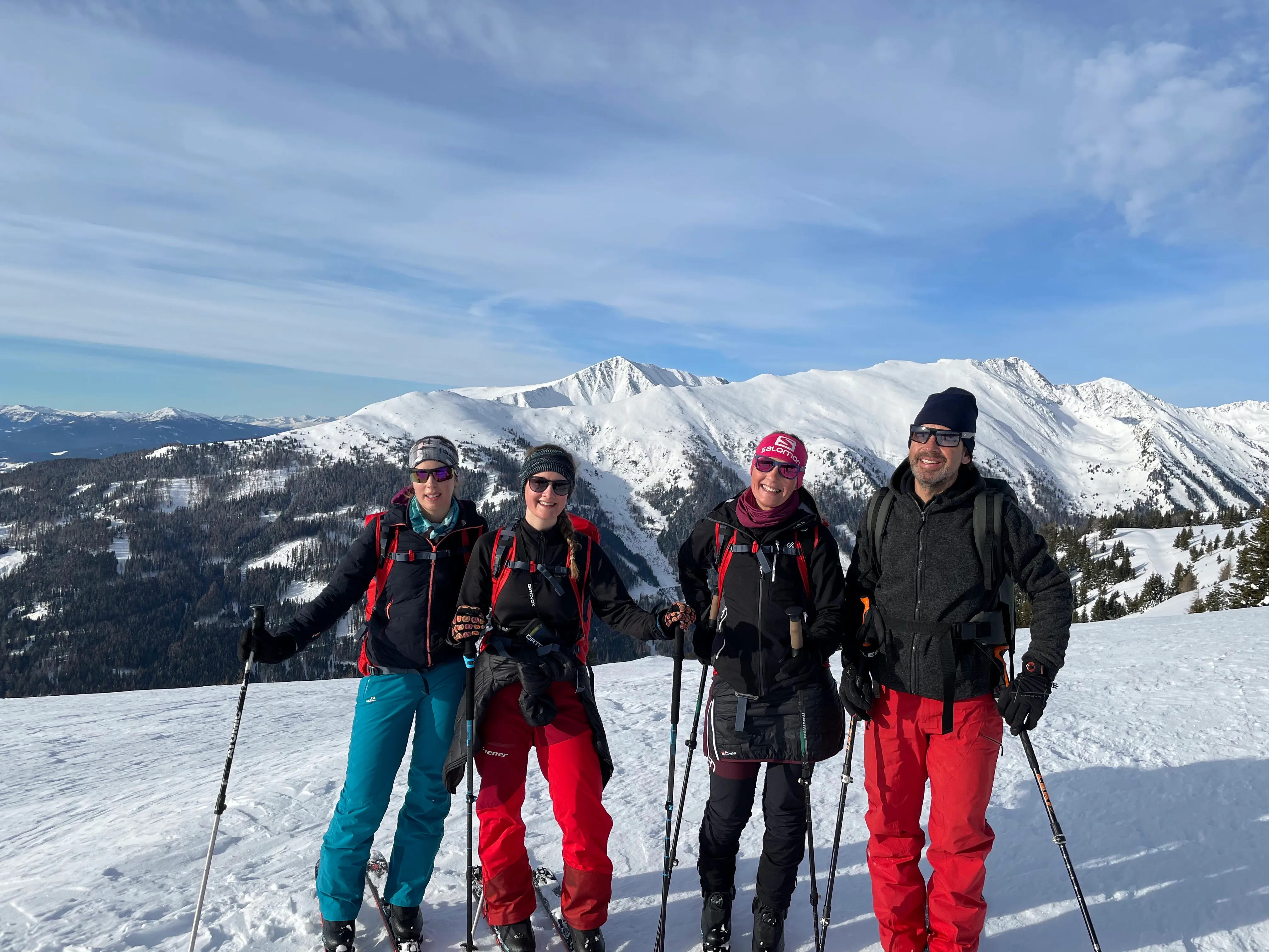 Vier Personen in Wintersportkleidung stehen mit Skistöcken auf einer verschneiten Berglandschaft mit schneebedeckten Bergen und blauem Himmel im Hintergrund.