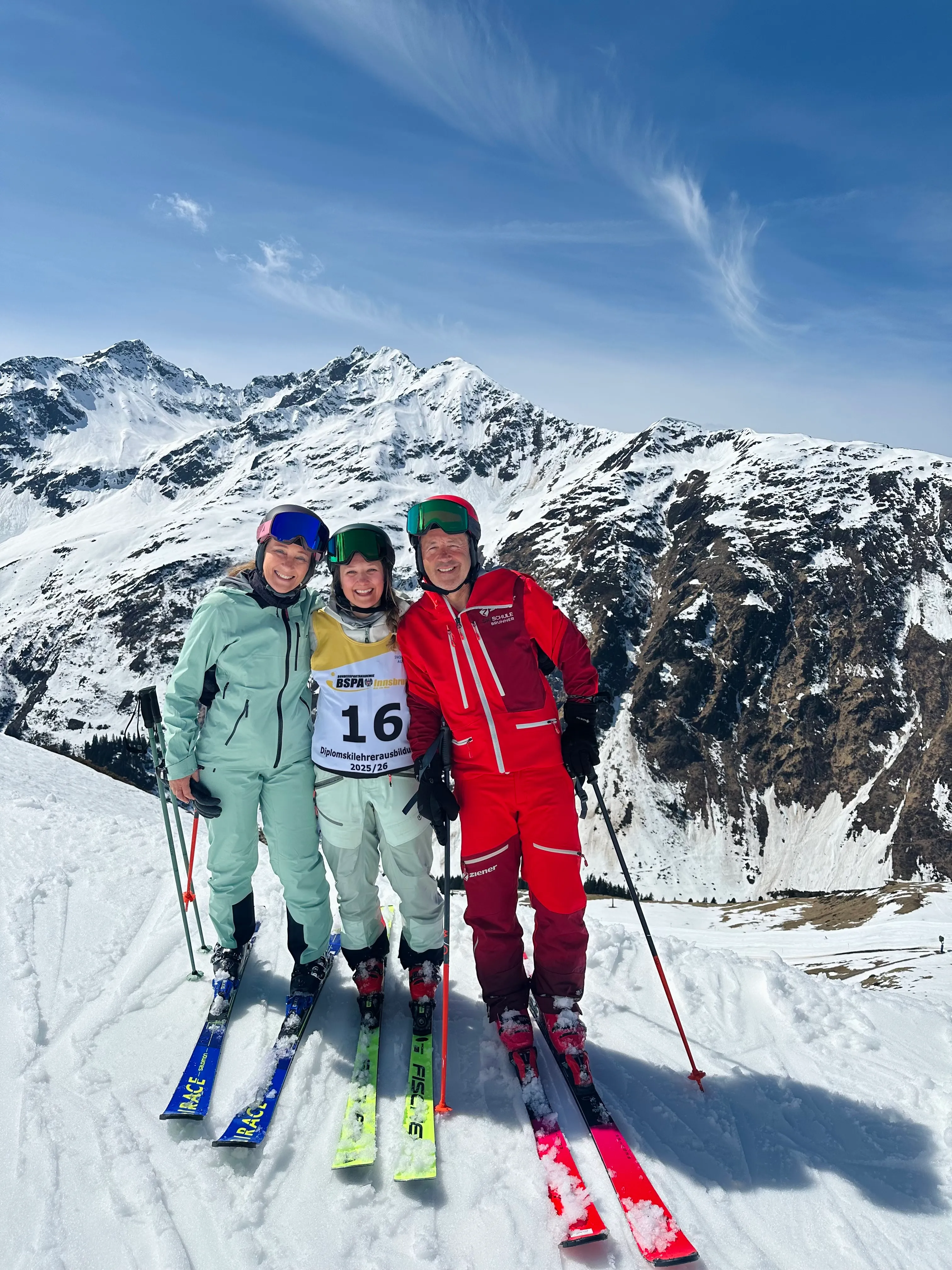 Drei Skifahrer posieren auf einer schneebedeckten Bergspitze bei sonnigem Himmel, umgeben von schneebedeckten Gipfeln; zwei Personen tragen hellblaue Skianzüge und eine Person einen roten Anzug, alle mit Skibrillen und Skistöcken.