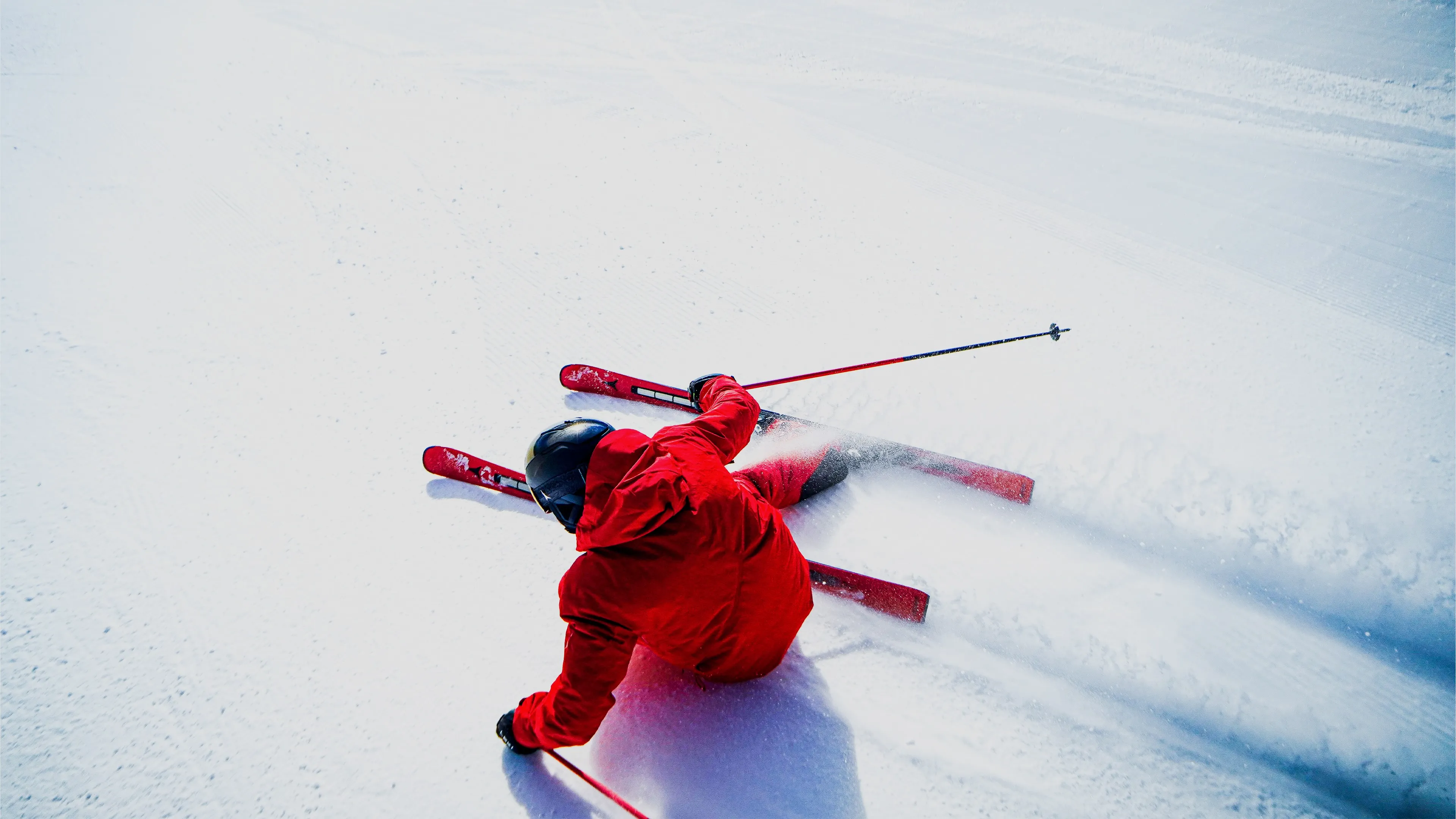 Skifahrer in roter Jacke und Helm fährt schnell im Pulverschnee, mit Skistöcken in der Hand, Schnee spritzt seitlich auf der Piste.