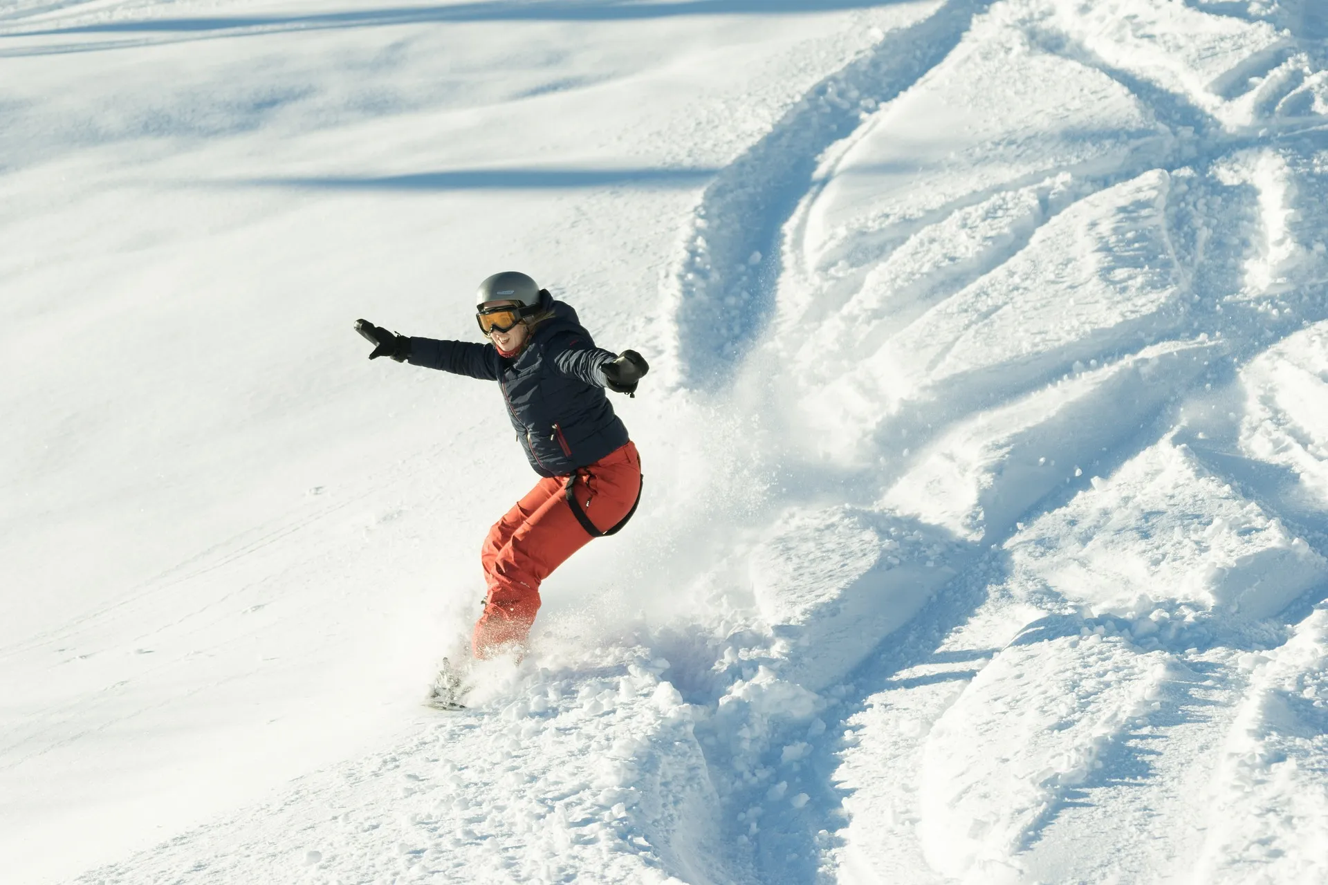 Eine Person in schwarzer Jacke, orangefarbener Hose, Helm und Schutzbrille fährt im frischen Schnee Snowboard und erzeugt dabei Schneestaub.