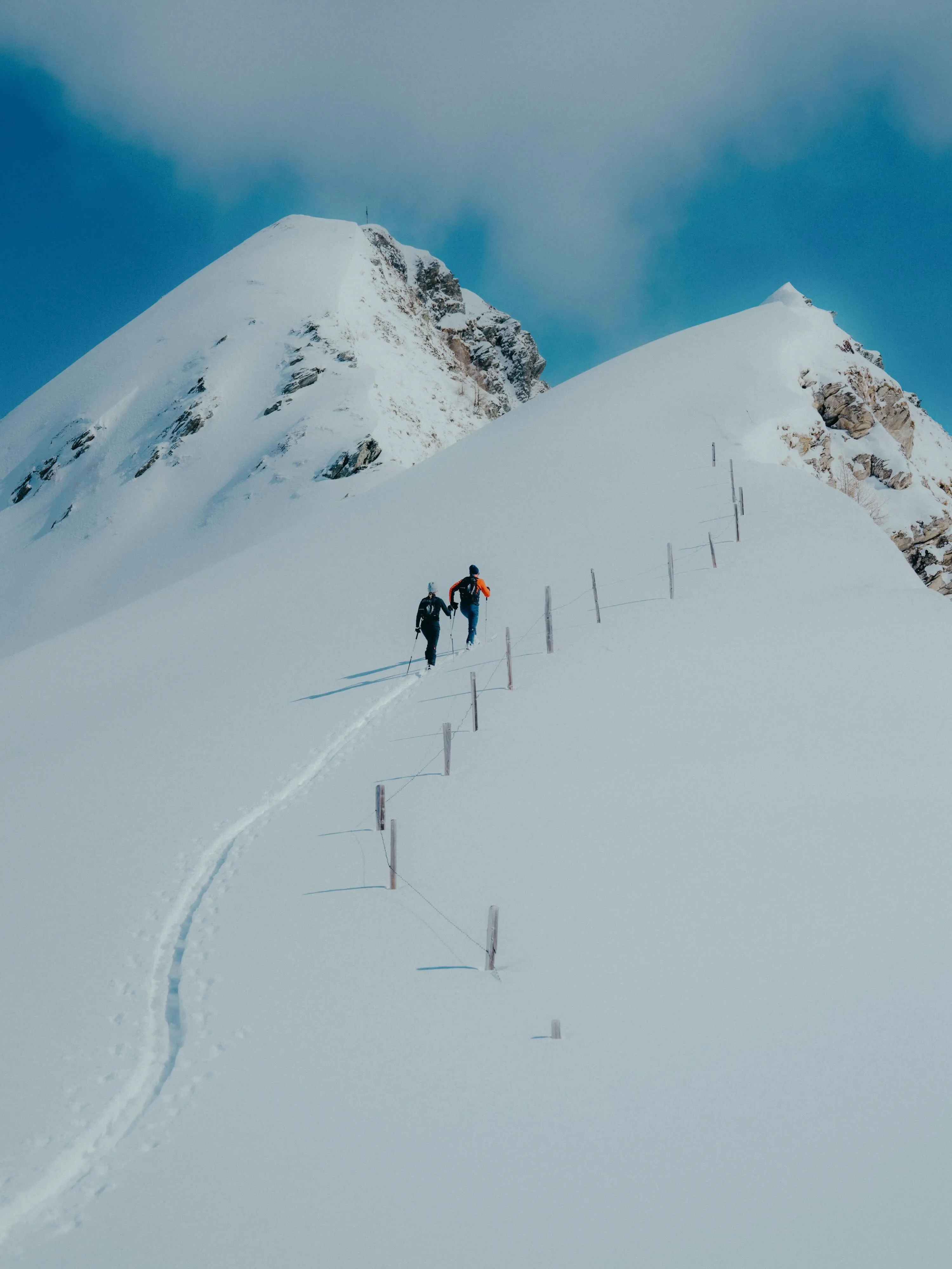 Zwei Skitourengeher steigen auf einer schneebedeckten Bergkuppe bei klarem Himmel auf, entlang eines Zauns mit Holzpfosten.