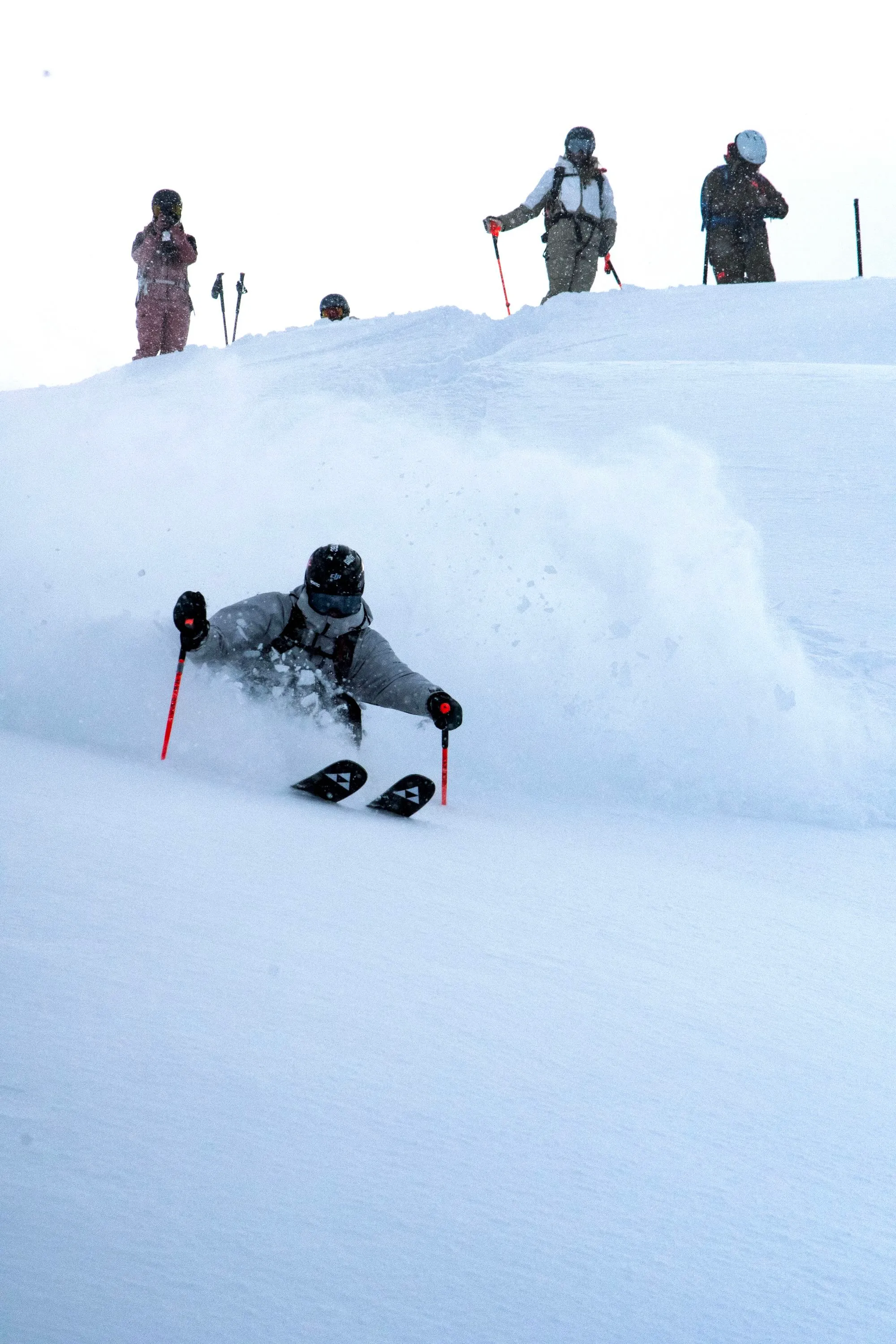 Ein Skifahrer fährt eine steile, unberührte Schneepiste hinab und wirbelt dabei frischen Pulverschnee auf, während drei Beobachter oben am Rand des Abhangs stehen und zuschauen.
