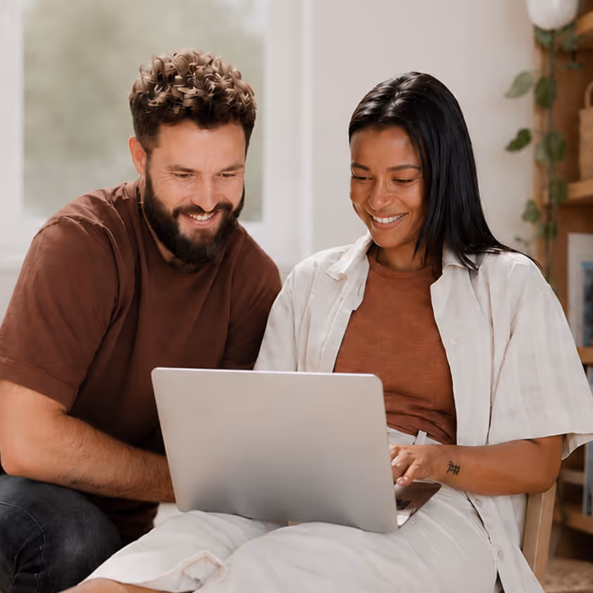Smiling man and woman sitting indoors, looking at a laptop together.