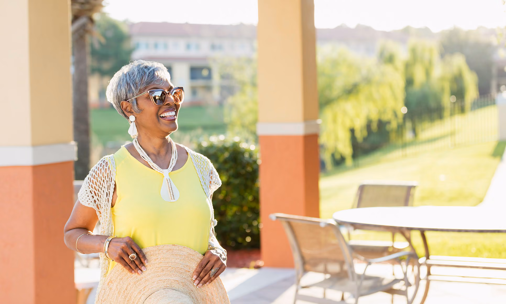 Smiling older woman in yellow shirt holding a straw hat, wearing sunglasses outdoors on a sunny day.