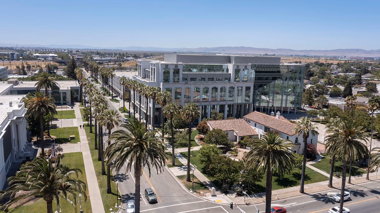 Aerial view of a modern glass office building, palm trees lining streets, and smaller houses nearby.
