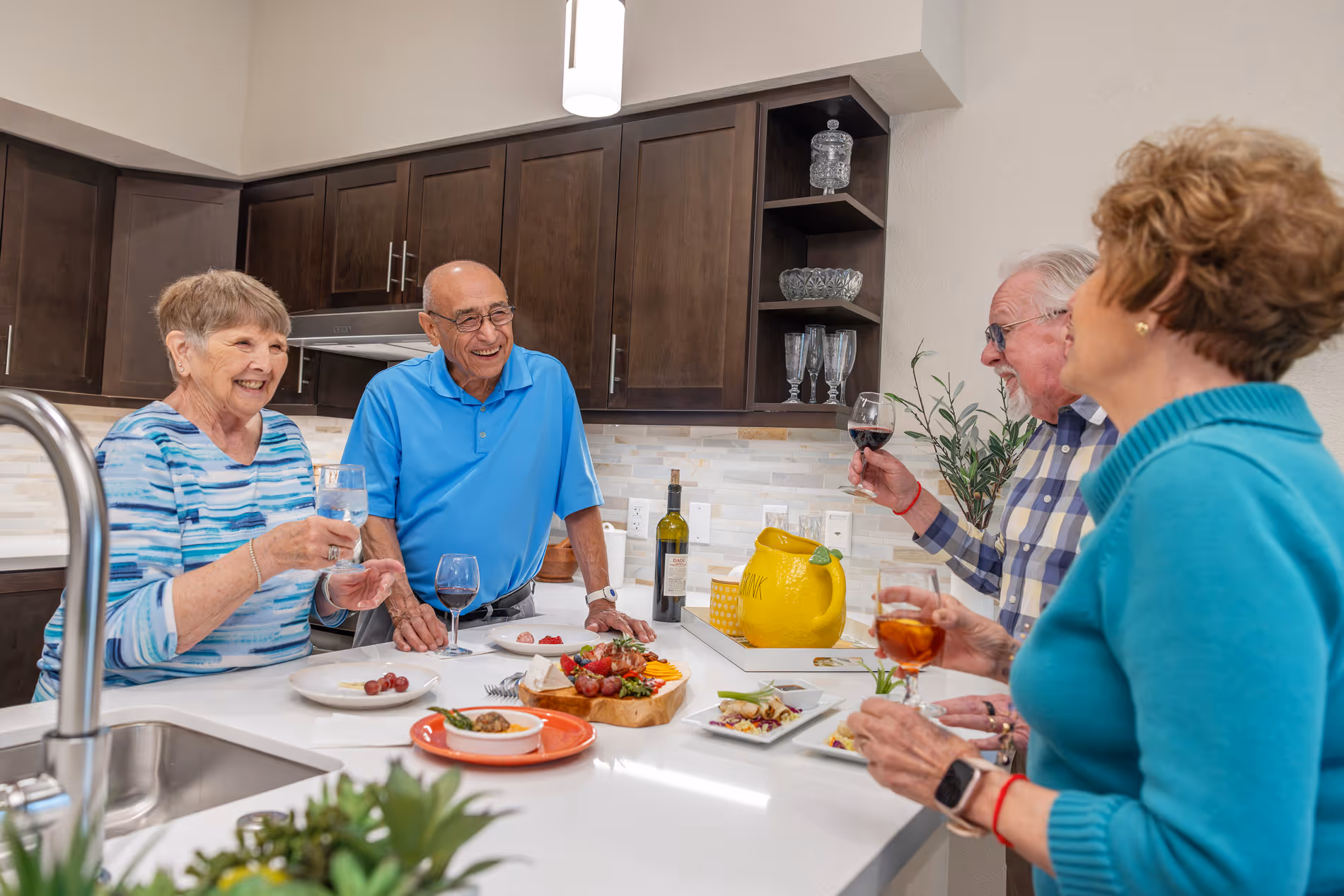 Four seniors enjoying drinks and appetizers together in a modern kitchen with dark cabinets.