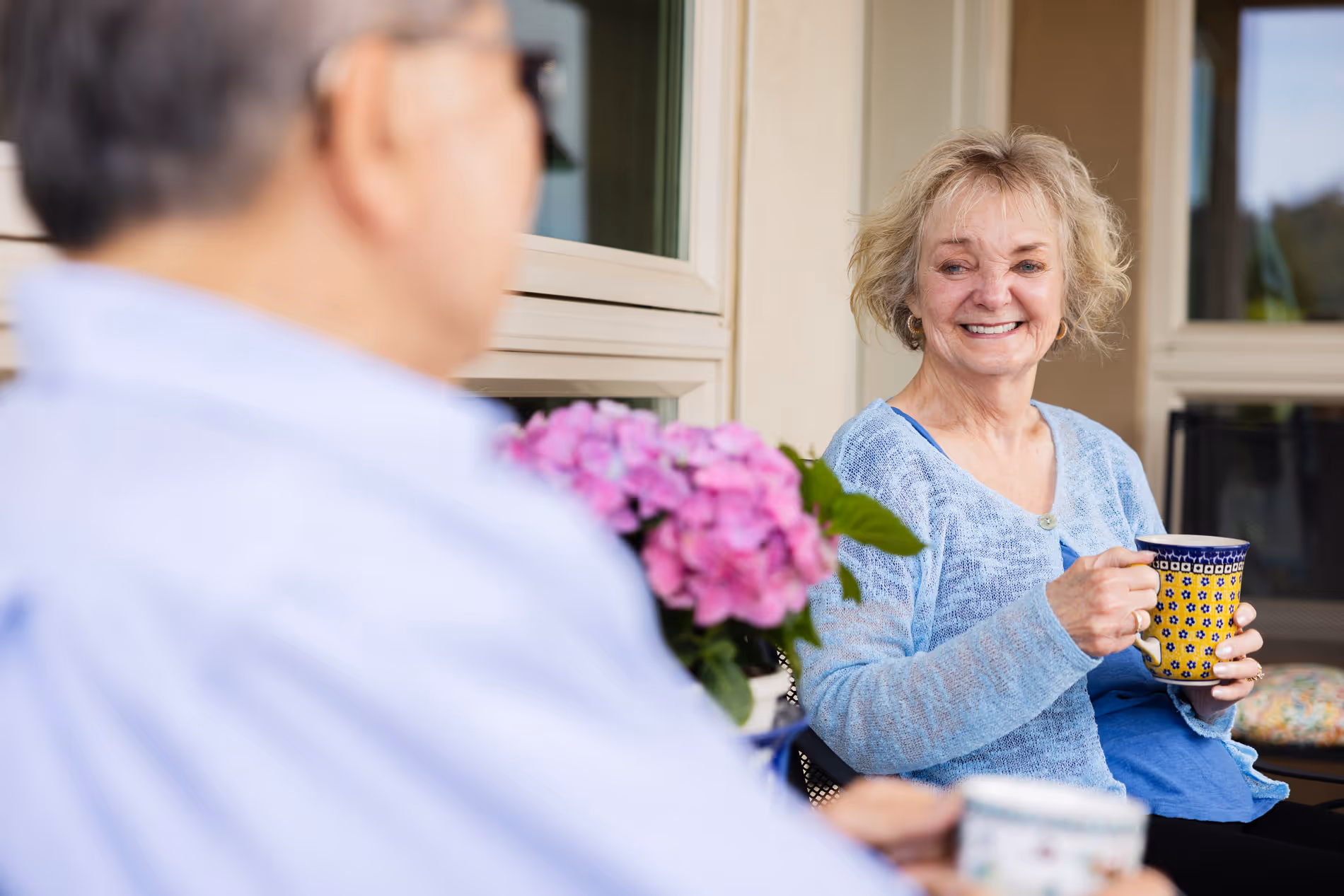 Smiling older woman holding a yellow mug, sitting outdoors with a man holding a cup nearby.