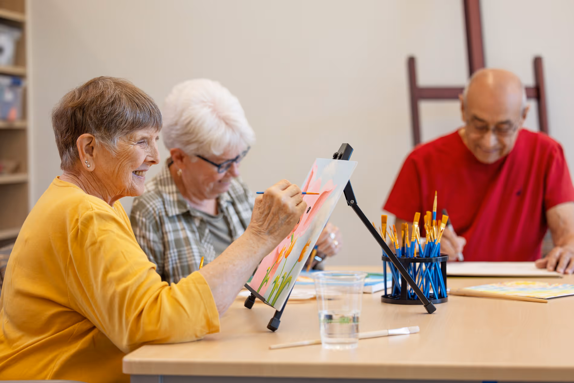 Three seniors painting at a table with brushes and canvases, smiling and focused on their work.
