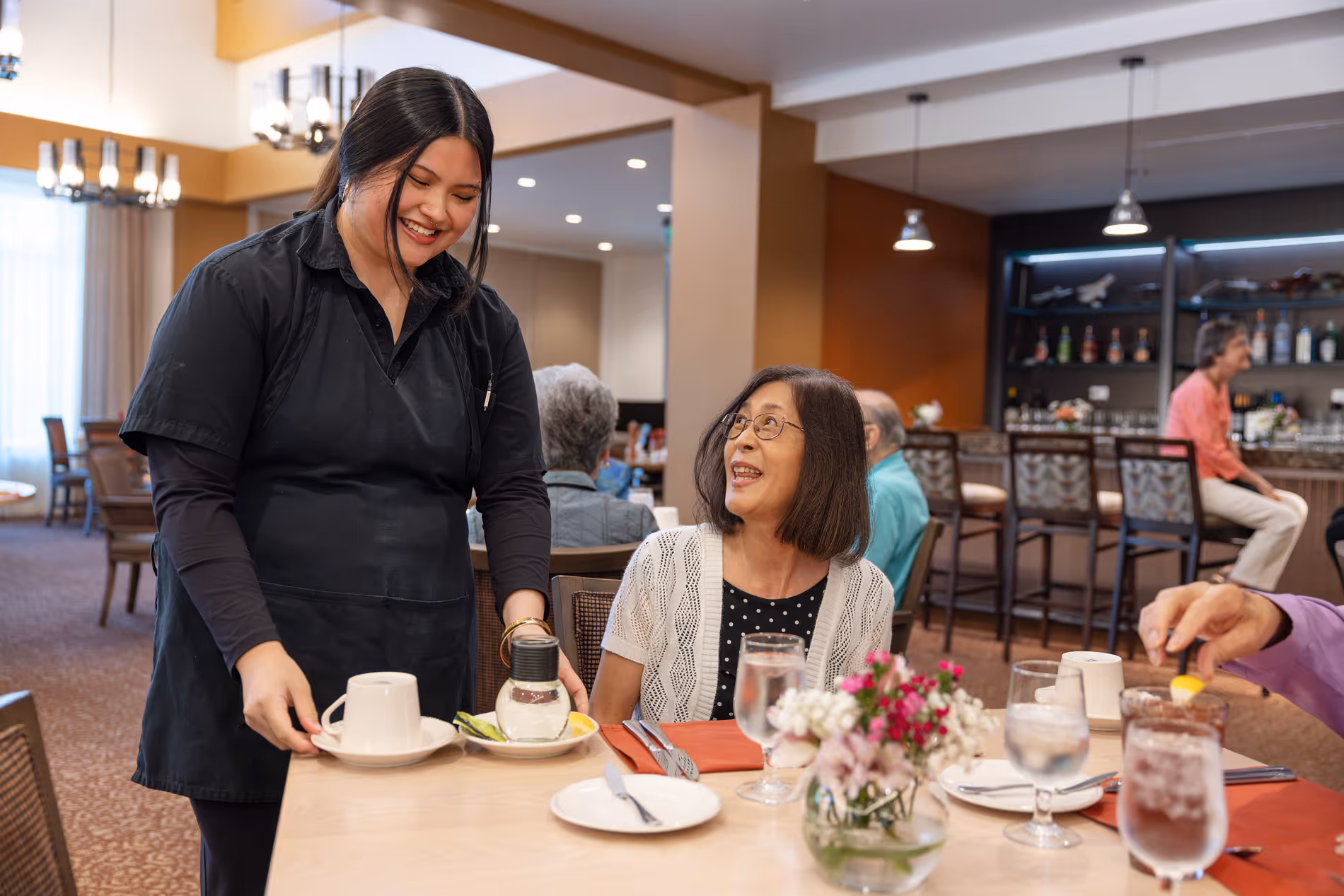 Smiling waitress serves a cup of tea to an elderly woman seated at a restaurant table with flowers.