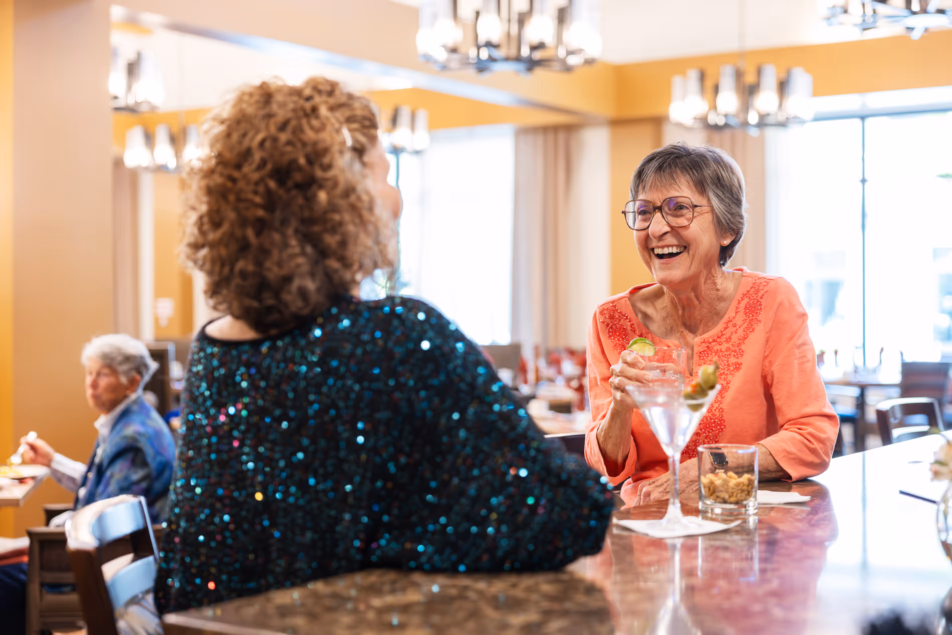 Two older women chat and smile over drinks at a restaurant bar with another woman dining in the background.