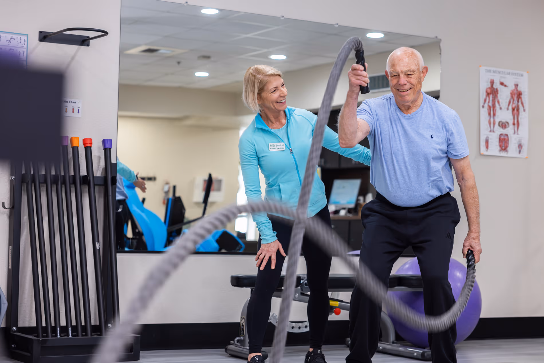 Older man using battle ropes guided by a female fitness trainer in a gym setting.