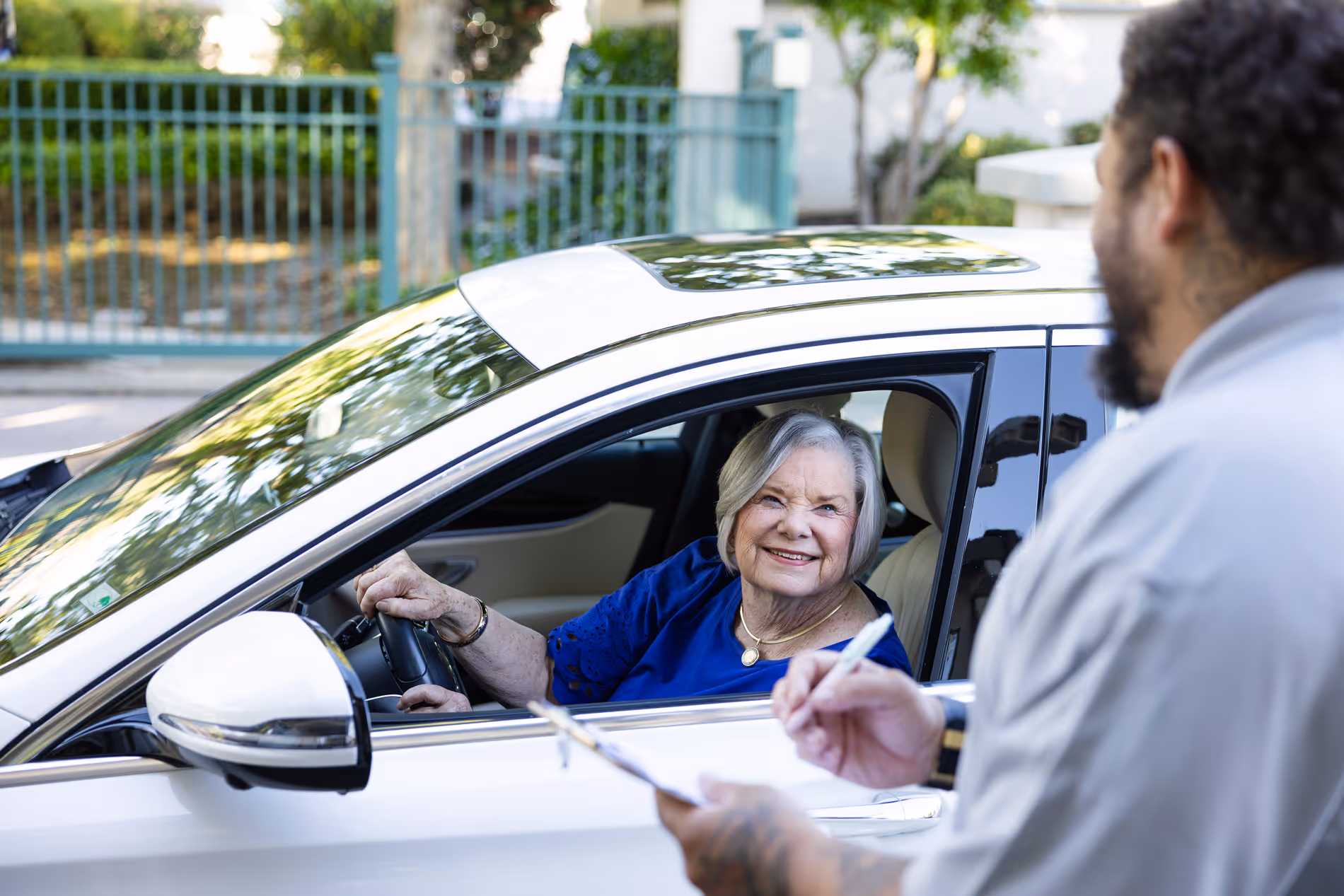 Senior woman smiling in a white car while talking to a man holding a clipboard outside.