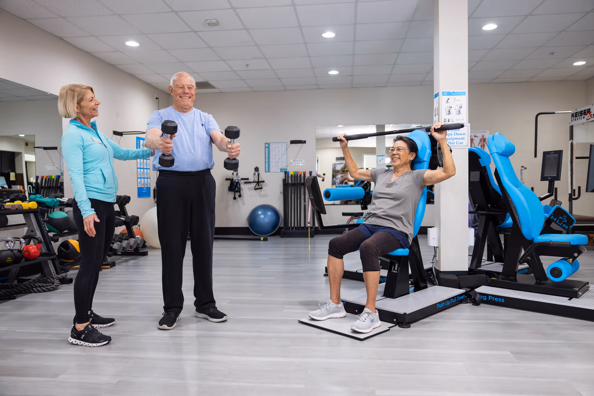 Older adults exercising with dumbbells and a machine in a bright gym with a female trainer.
