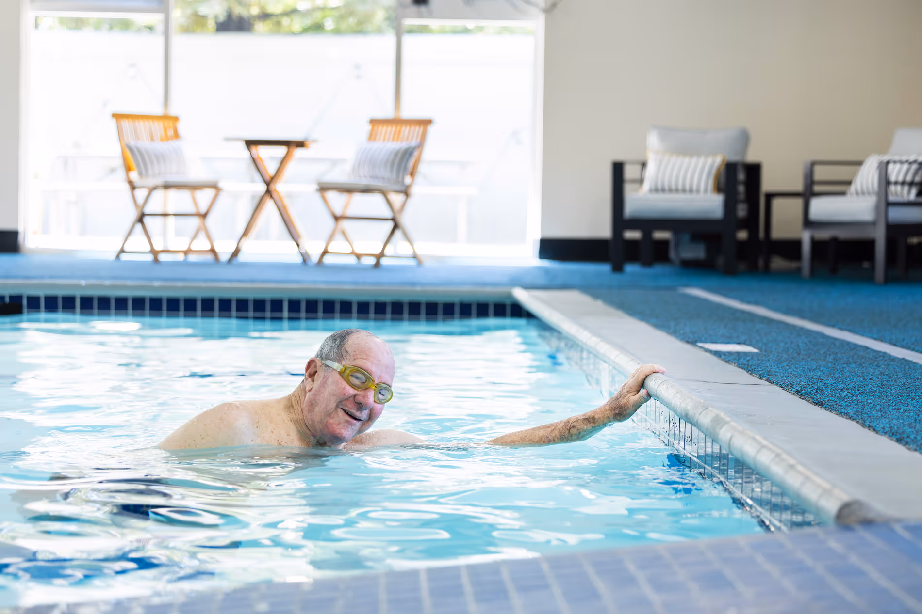Smiling elderly man wearing goggles swimming in an indoor pool holding the pool edge.