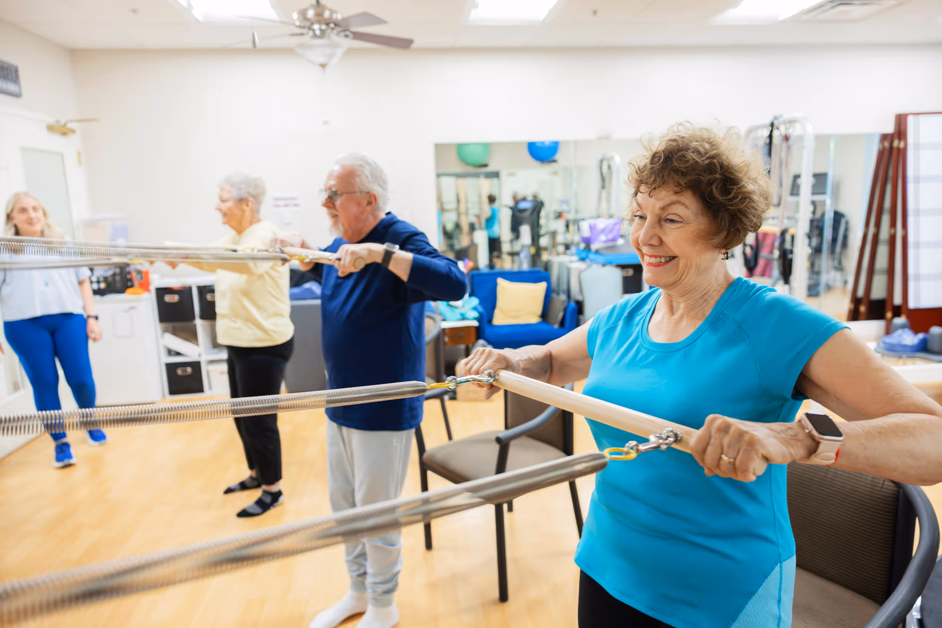 Three seniors do seated resistance band exercises in a bright, well-equipped fitness room.