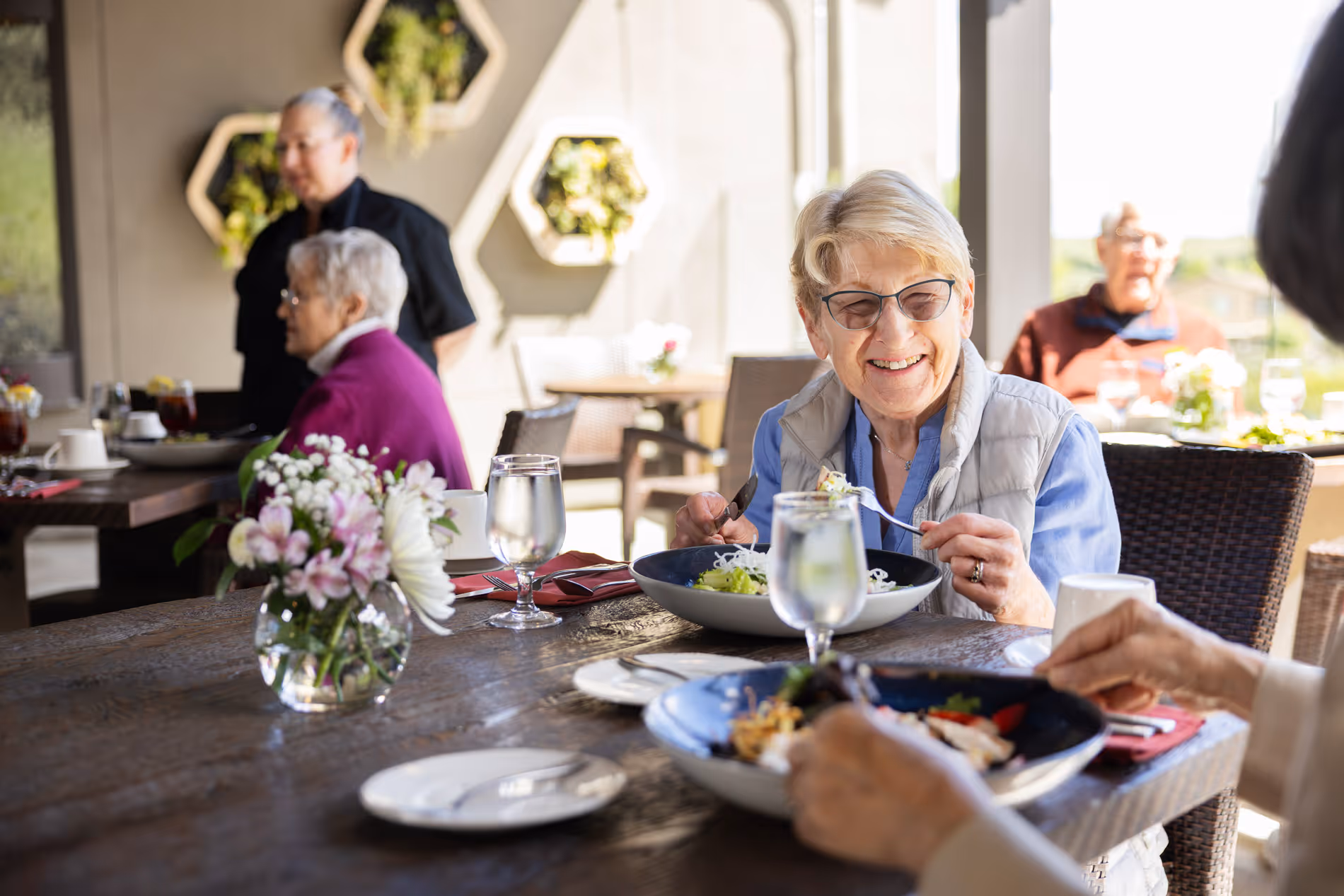 Smiling elderly woman with glasses eating salad at a table with others and flowers in a vase.