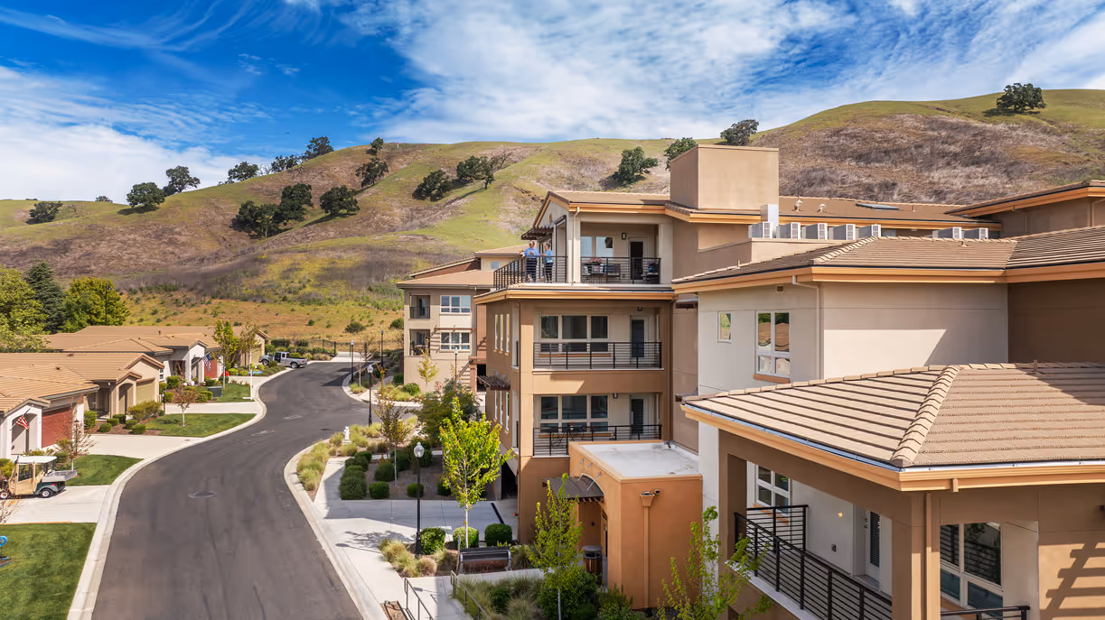 Residential buildings along a curved road with green hills and a blue sky in the background.